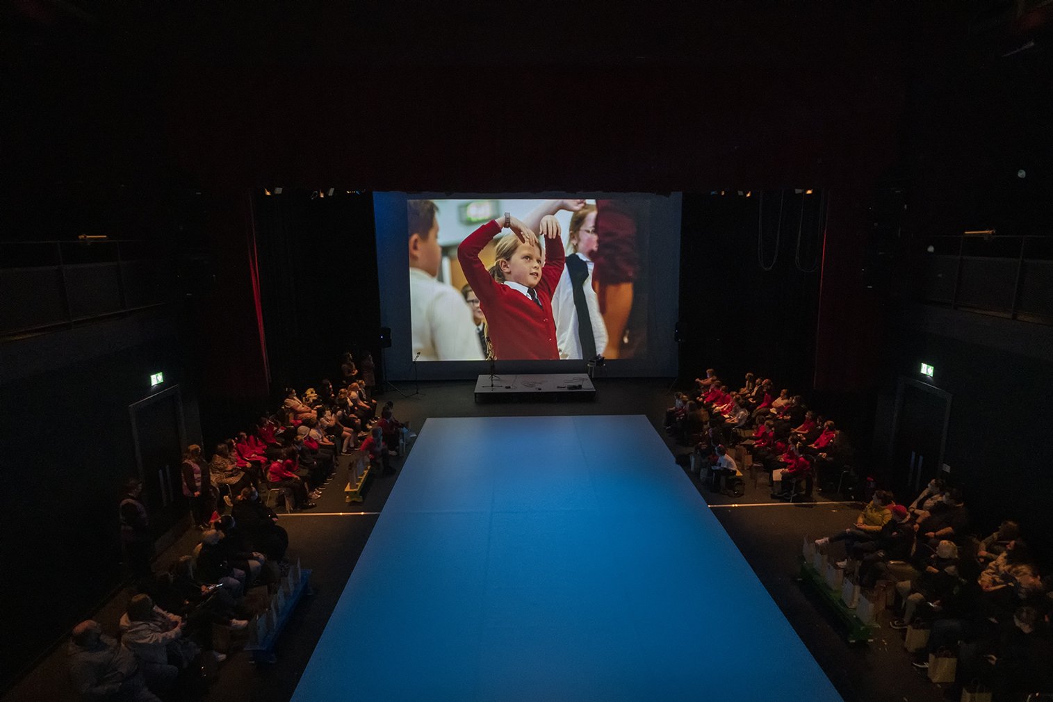  Zoomed out shot of a hall with a big screen showing photos to the audience sitting at the sides of the room and a cleared space along the middle of the room 