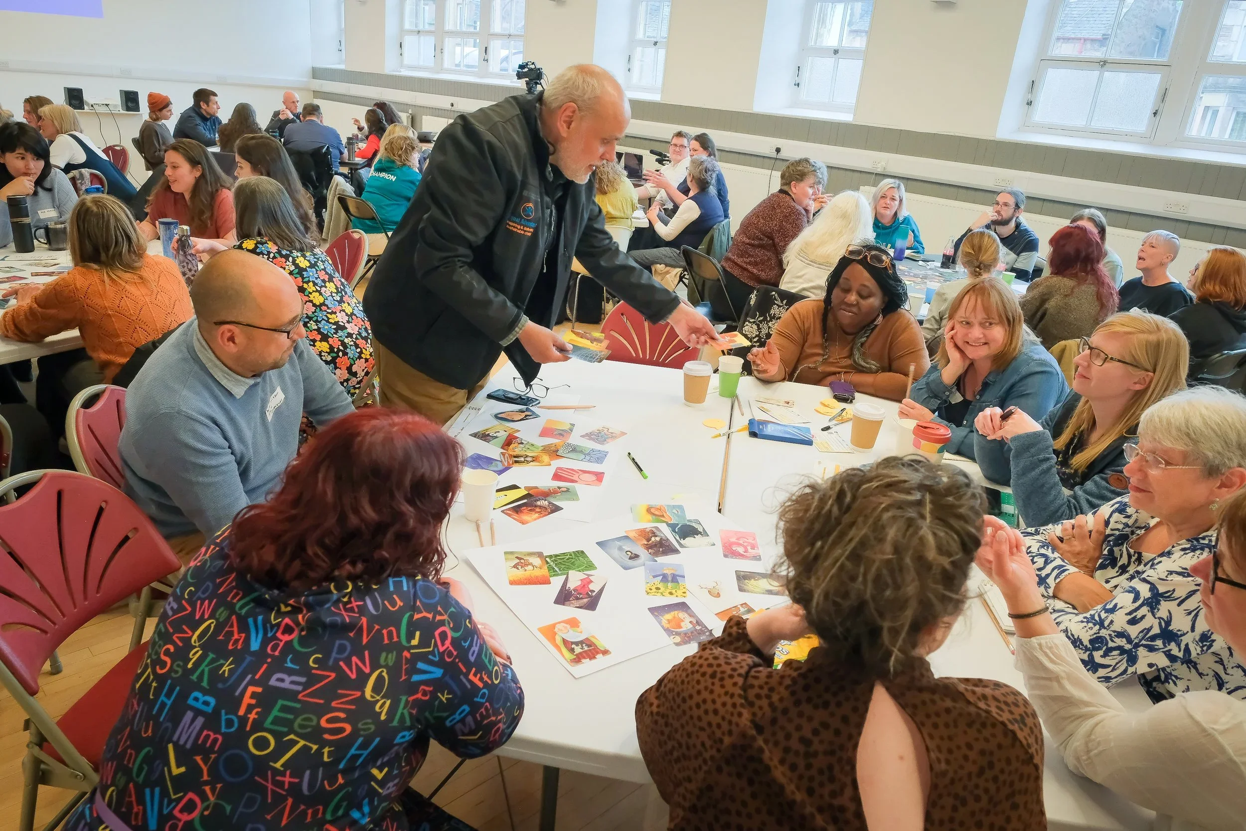 Participants at the 2024 Community Knowledge Matters Gathering in Inverness engaged in a group discussion around a table. They are looking at images and talking animatedly, illustrating a collaborative and reflective environment.