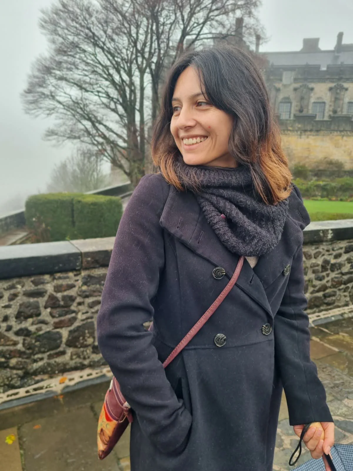 Natalia, outdoors, smiling in front of a historic building on a misty day