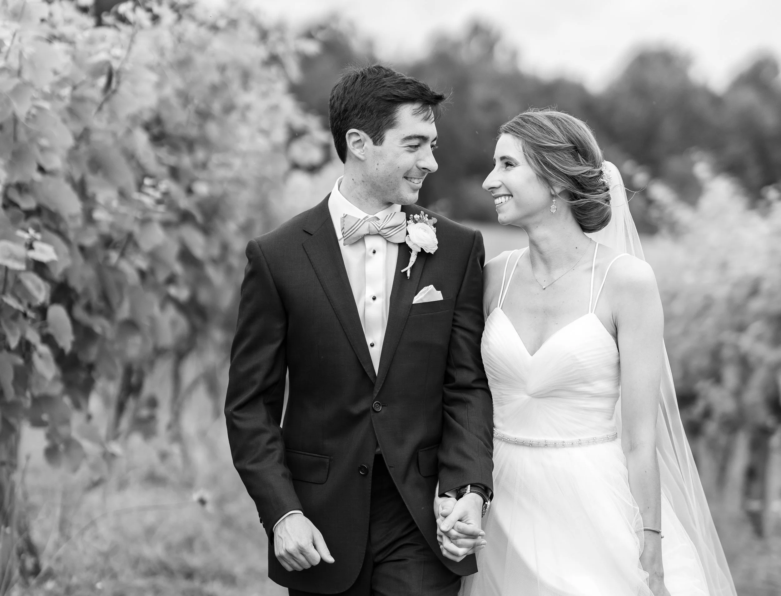 Black and white photo of bride and groom walking through a vineyard in Theia gown and J Crew suit
