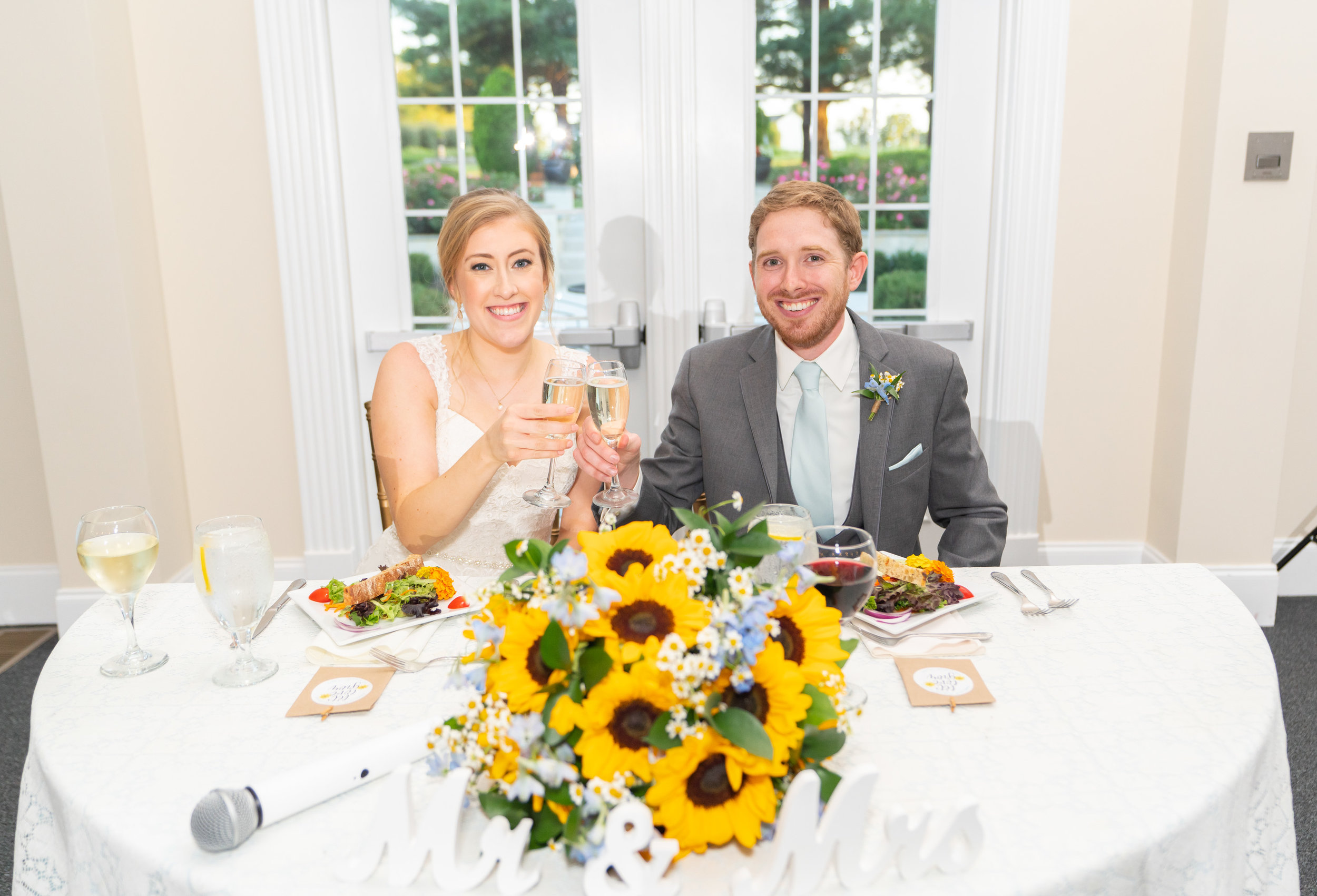 Bride and groom toast at sweetheart table with sunflower bouquet