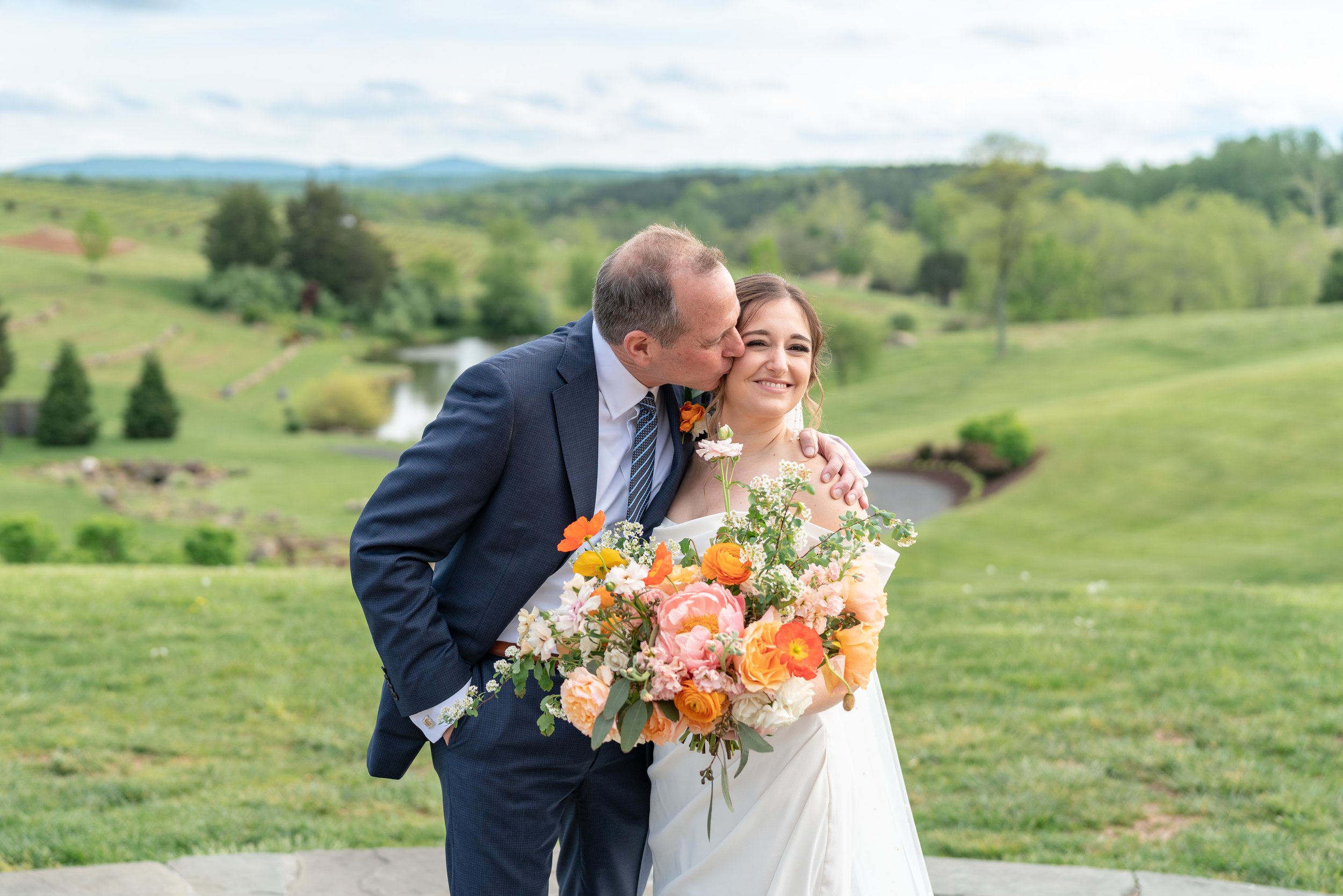 Bride's dad kisses her on the cheek with hillside views behind them
