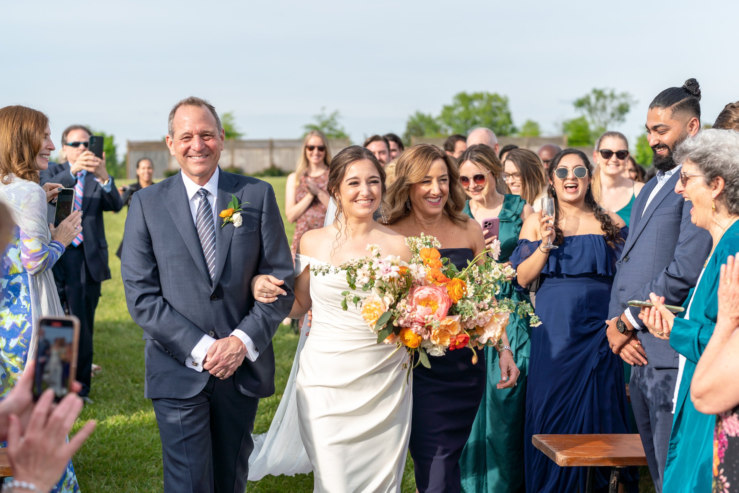 Bride walks with her parents and smiles at the groom under floral arch
