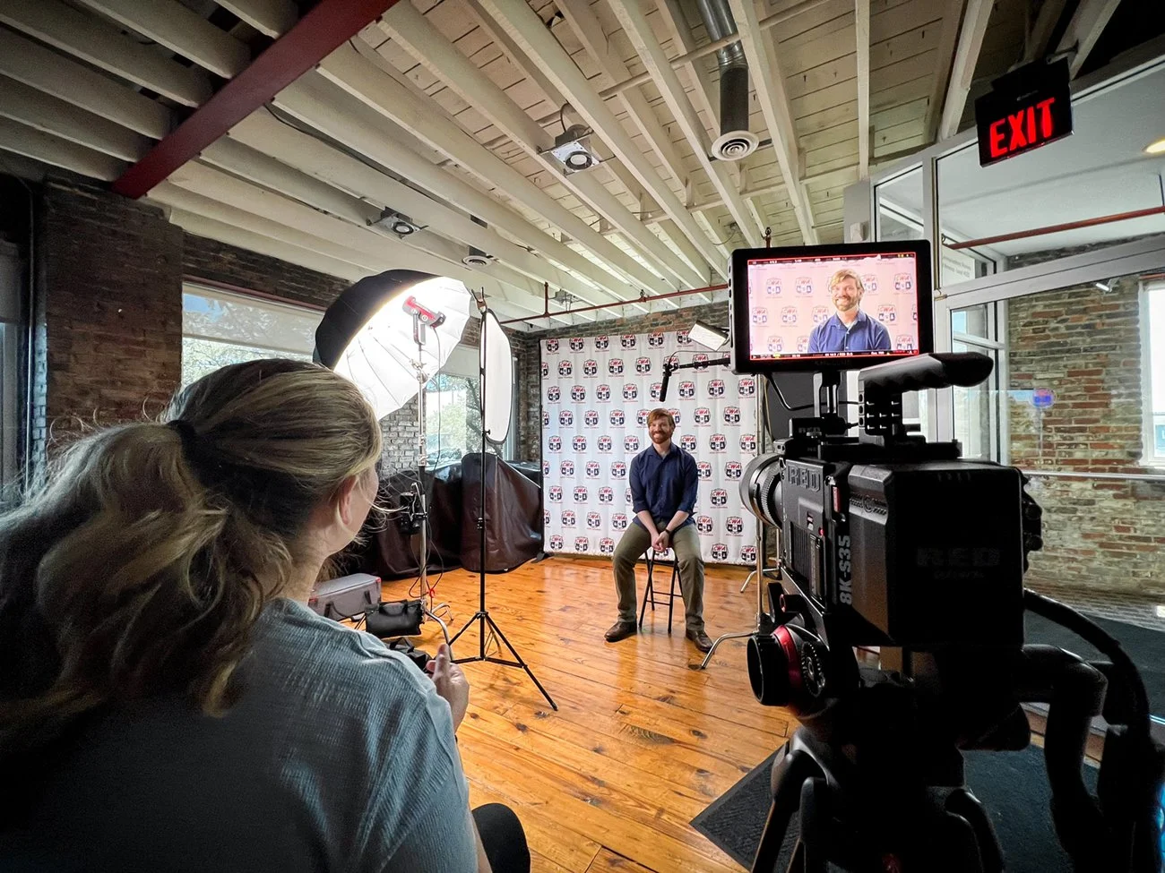 A man with a beard sits on a stool in front of a backdrop with logos, being filmed or photographed in an indoor studio with professional lighting and camera equipment, including a person operating a camera and a lighting umbrella.