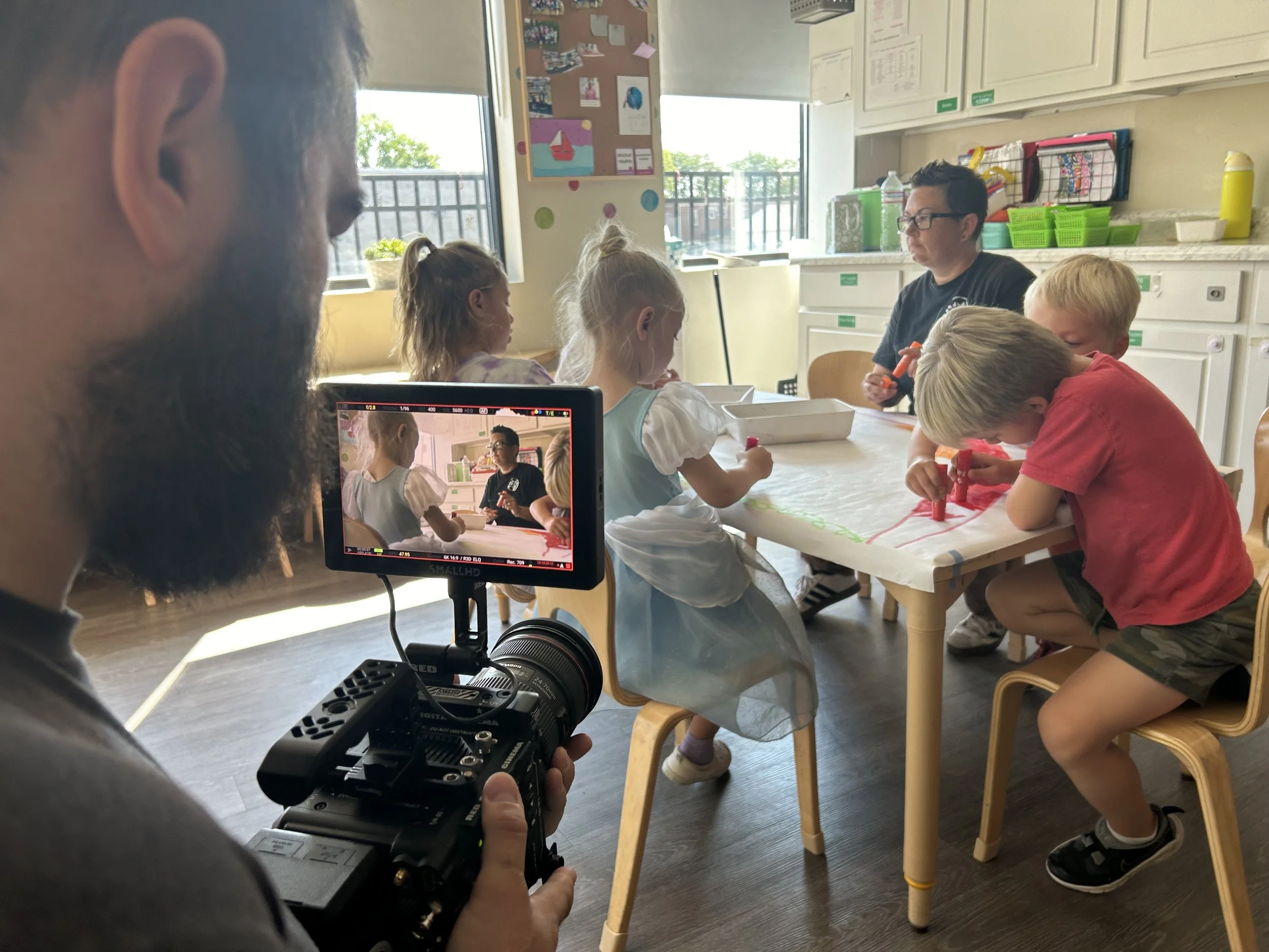 A man with a beard is filming children at a table in a classroom, with a focus on the camera's screen showing the children and a woman at the head of the table. The children are engaged in an art project.