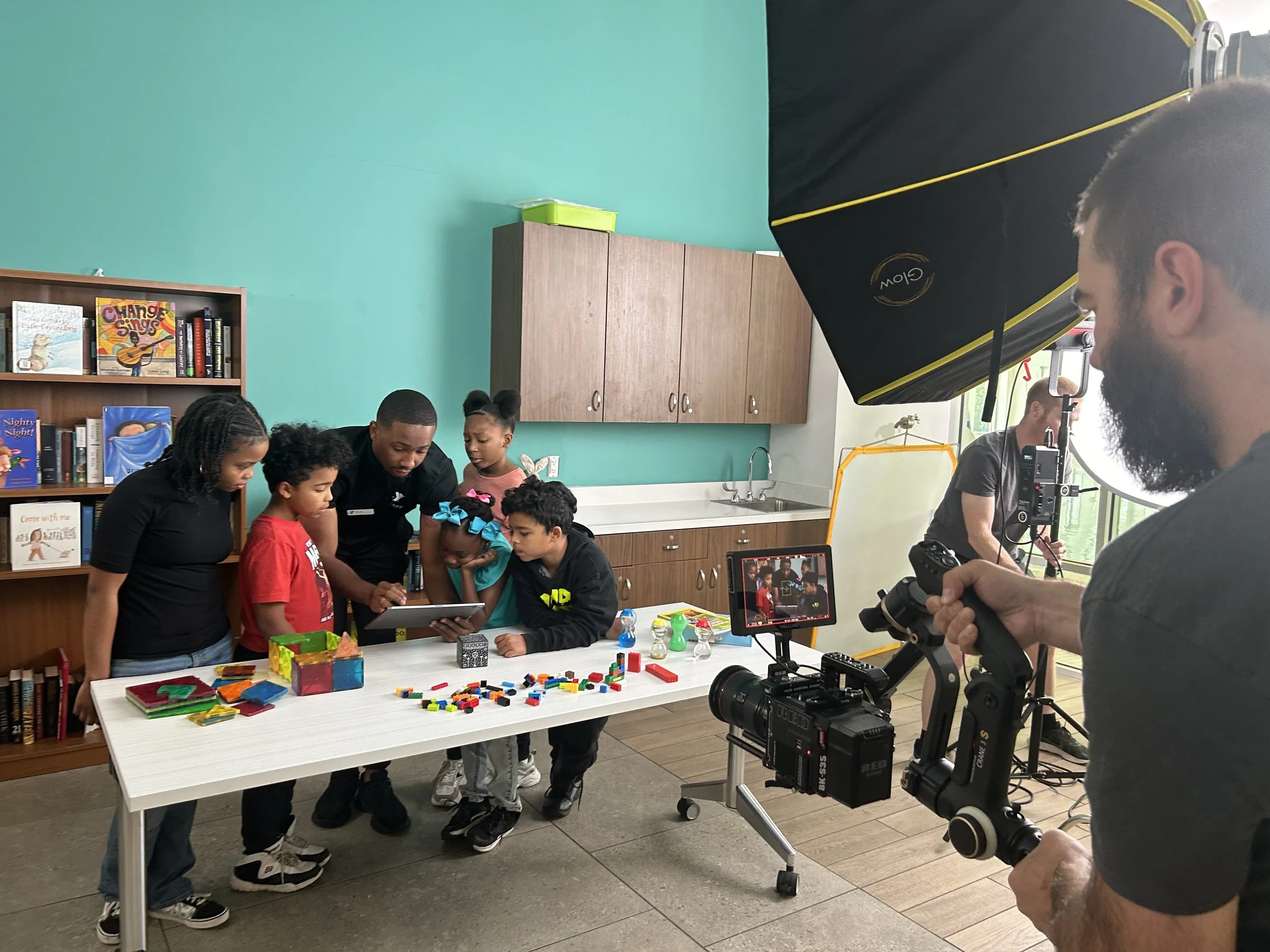 Filmmaker filming children gathered around a table with toys and books in a classroom.