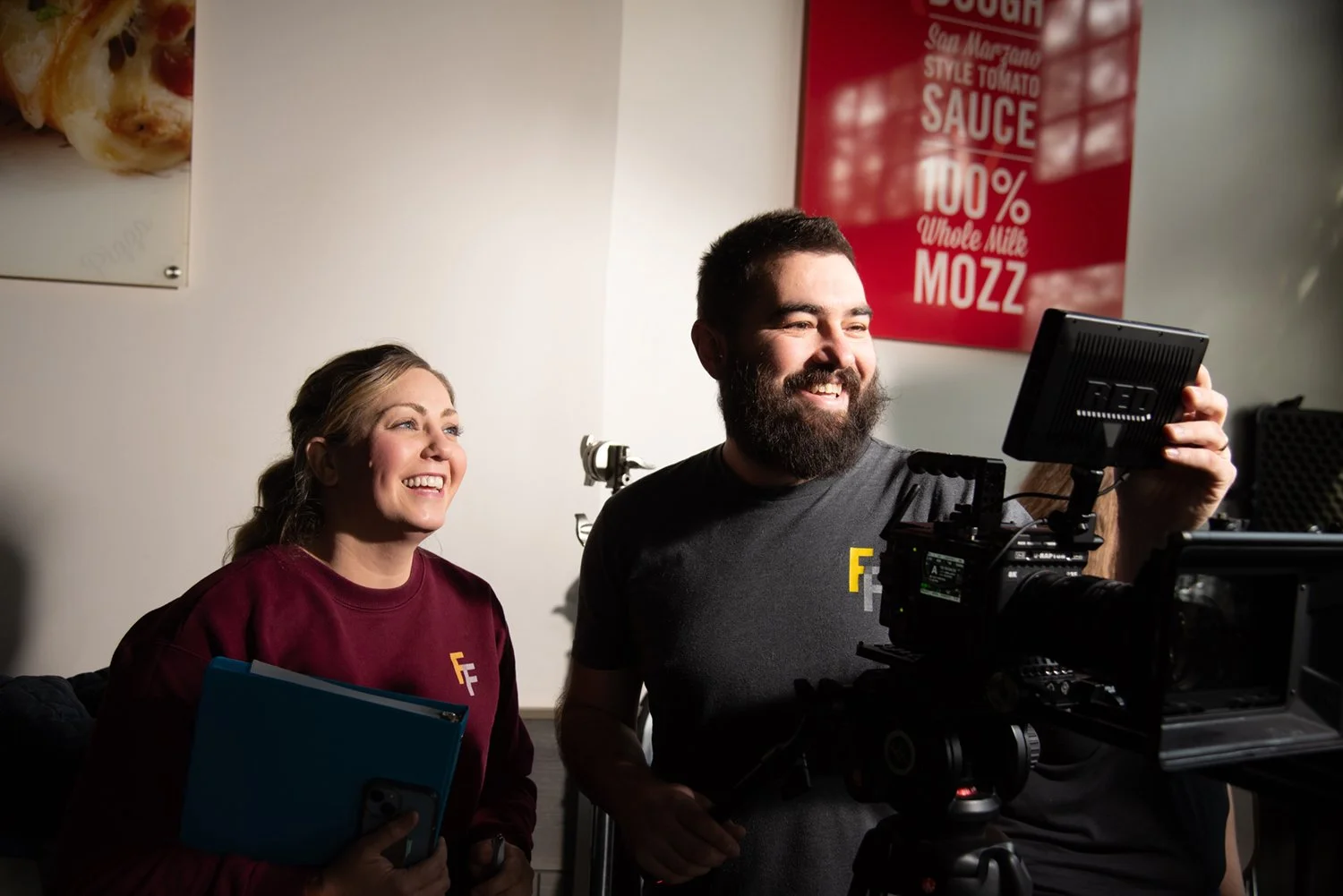 Two smiling people, a woman and a man, on a film or video production set. The woman holds a notebook and a phone, while the man operates a professional camera. They are indoors, with a white wall and a red poster in the background.