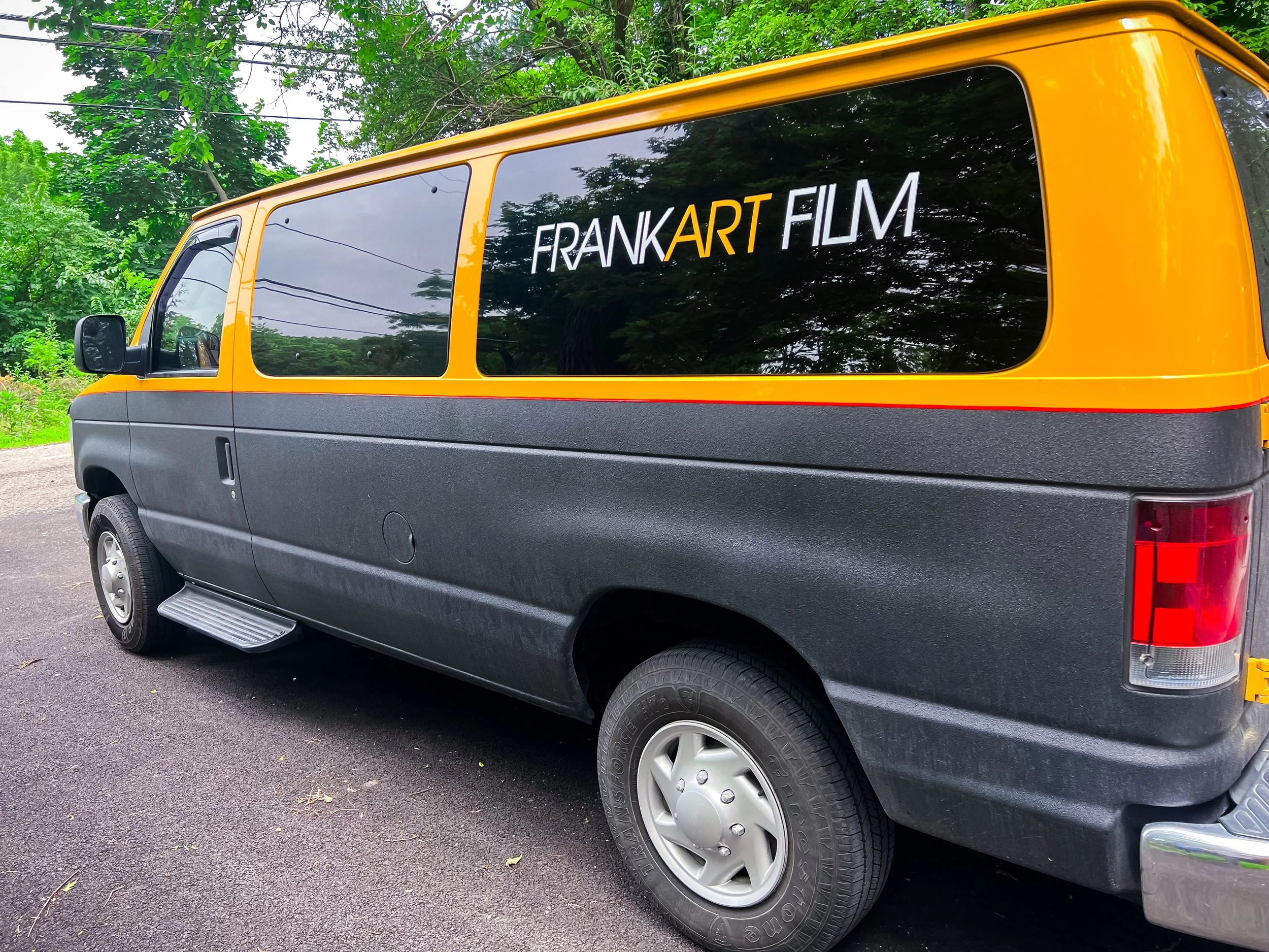 A yellow and black van parked on a paved surface with green trees in the background, featuring a decal on the window that reads 'FRANK ART FILM'.