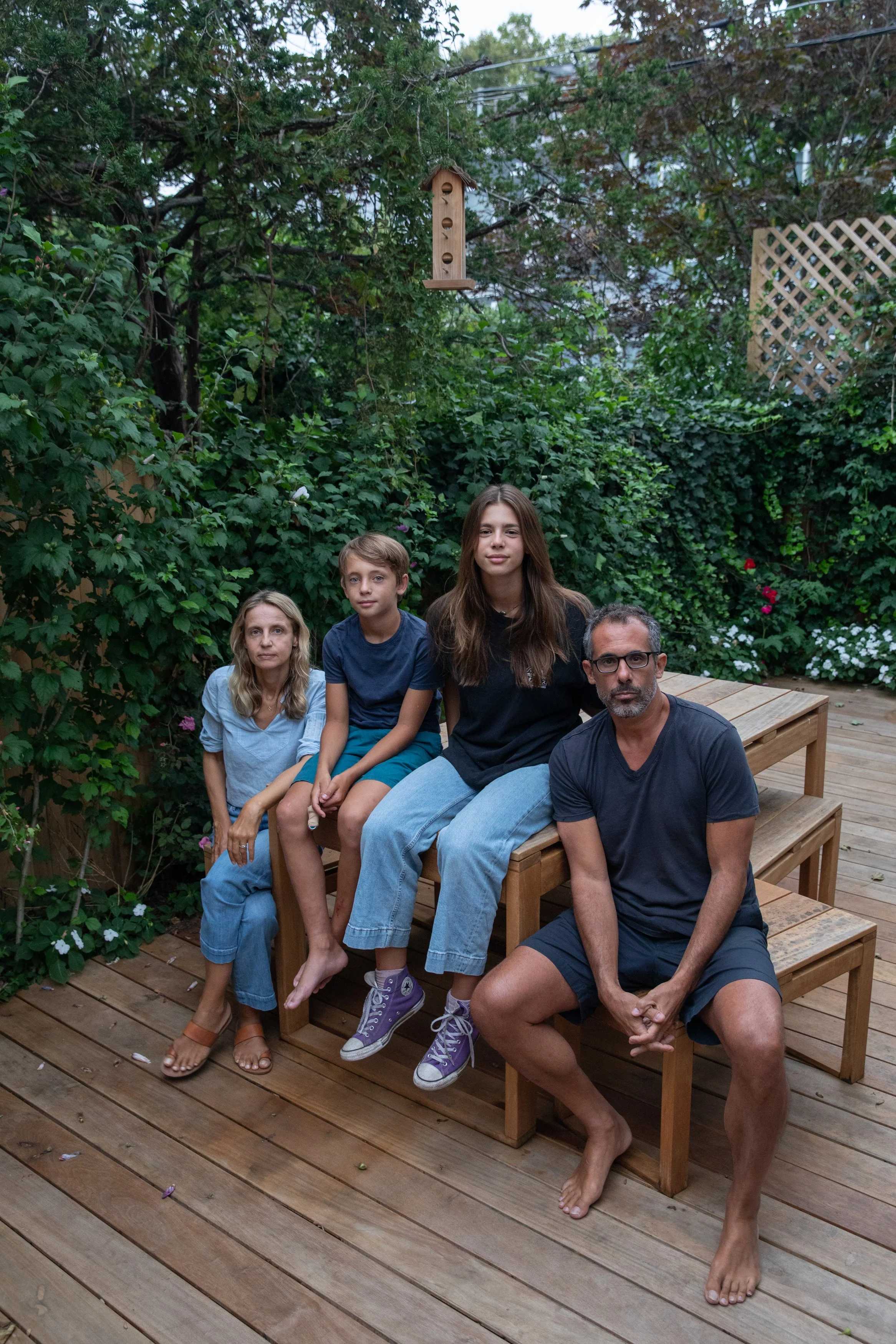 A family sitting on a wooden outdoor deck surrounded by lush greenery and flowers, with a birdhouse hanging from a tree overhead.
