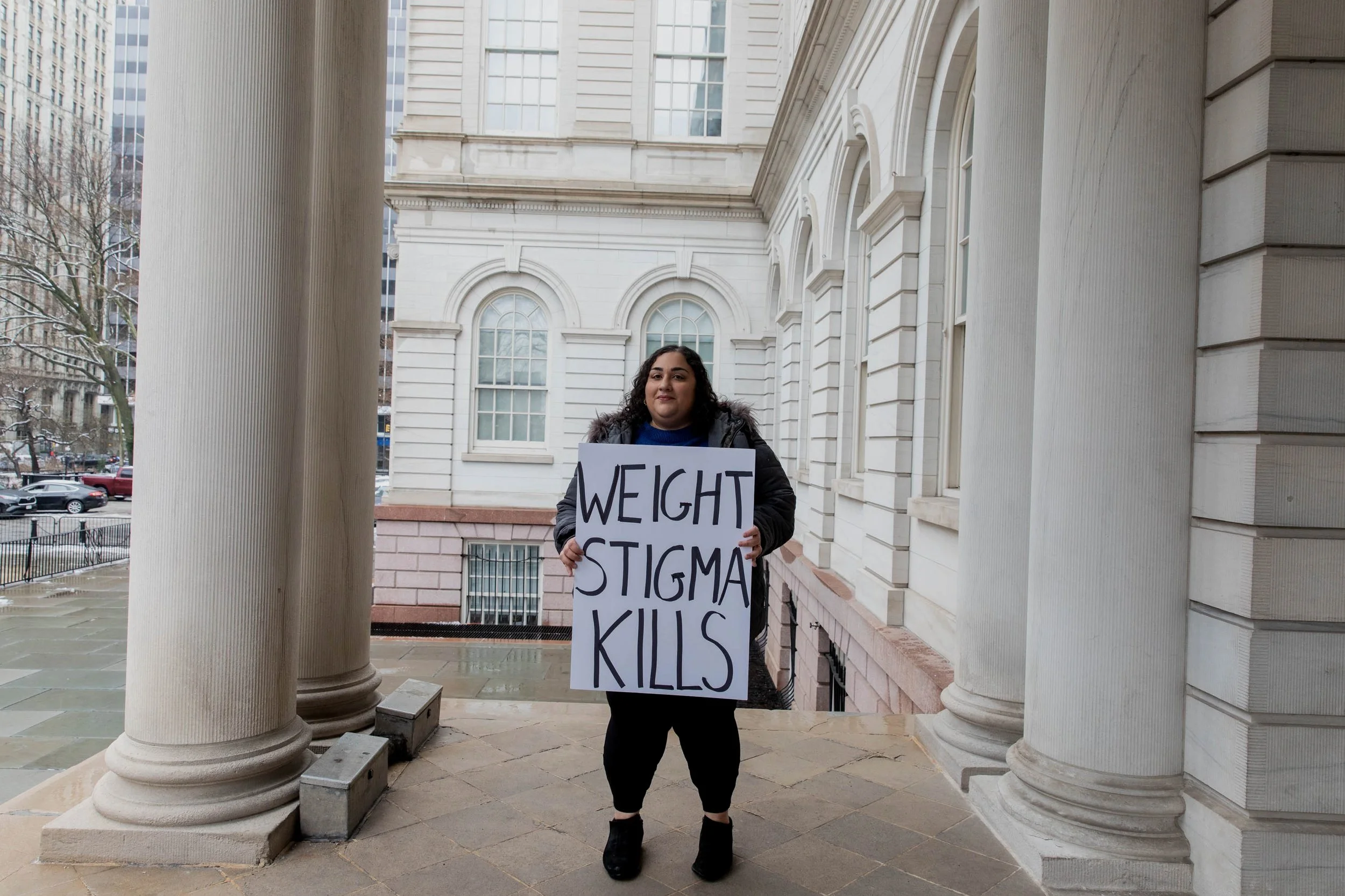 A woman holding a sign that reads 'WEIGHT STIGMA KILLS' standing between large classical columns in front of a historic building, with a city street visible in the background.