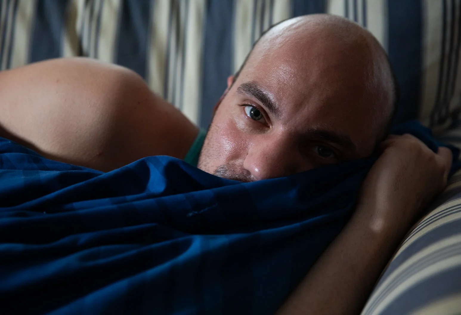 A man lying on a bed, resting his head on a pillow, with a striped pillowcase visible. He has a shaved head, is barefoot, and is looking directly at the camera with a relaxed expression.