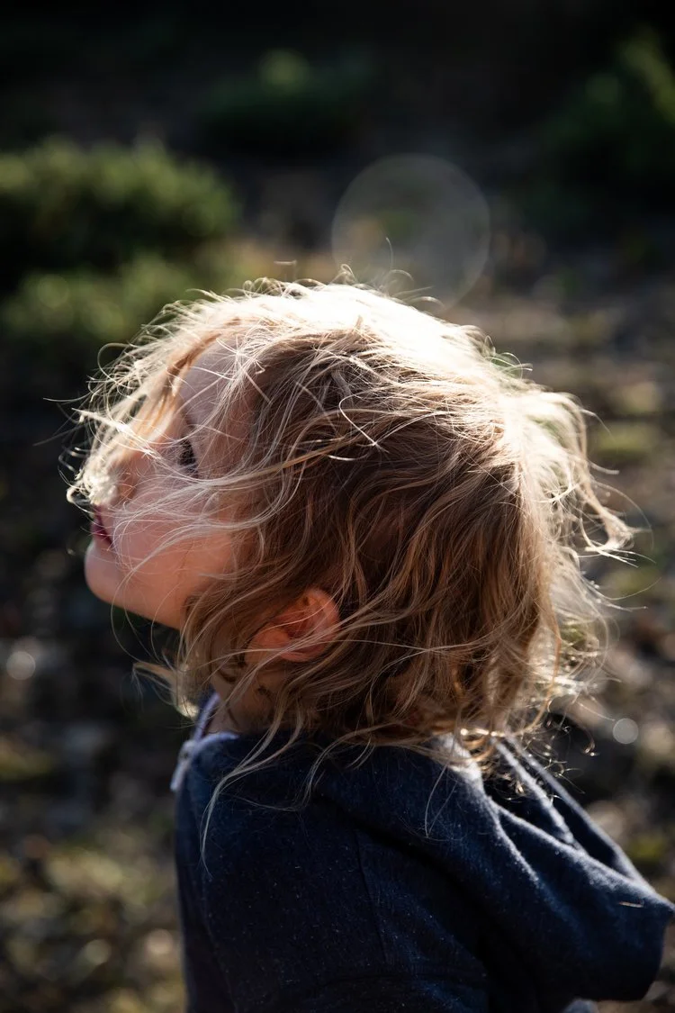 Side profile of a young girl with wavy blonde hair outdoors, with sunlight shining behind her.