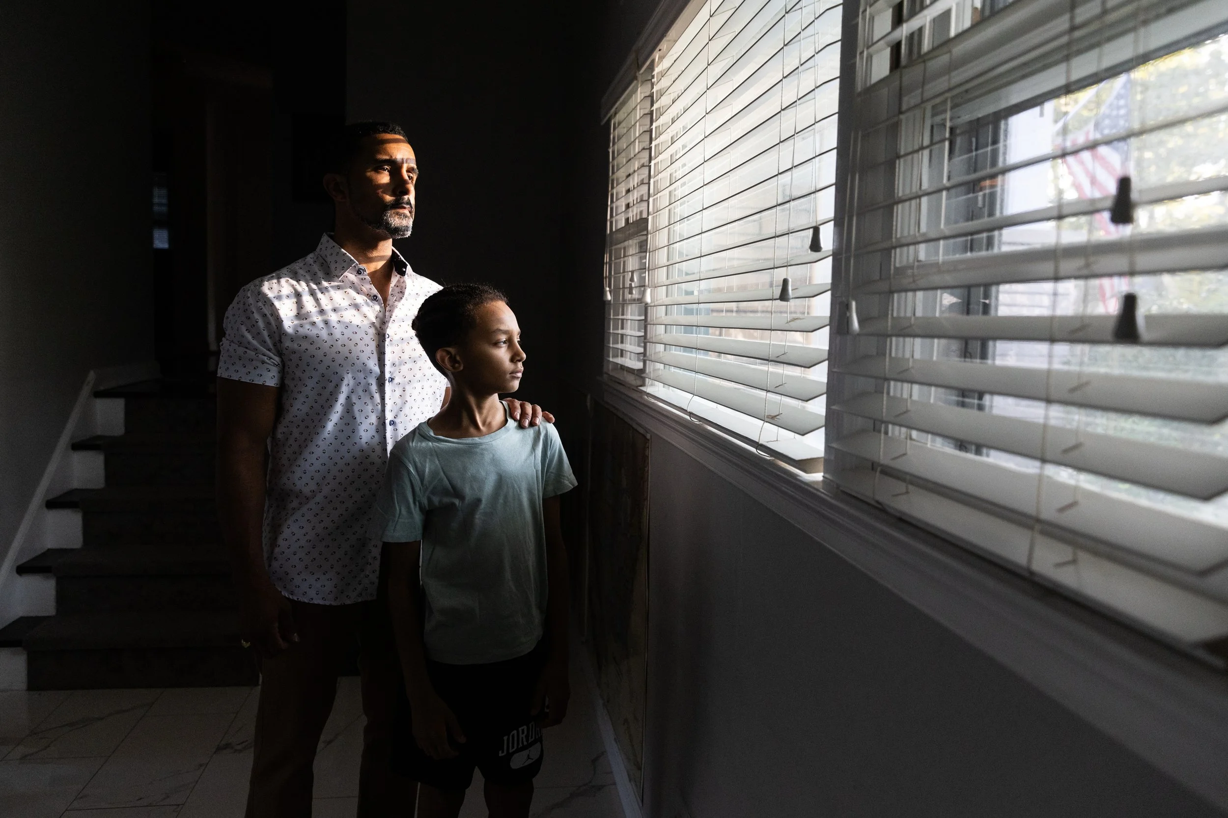 A man and a girl looking out a window with white blinds, lit by sunlight.