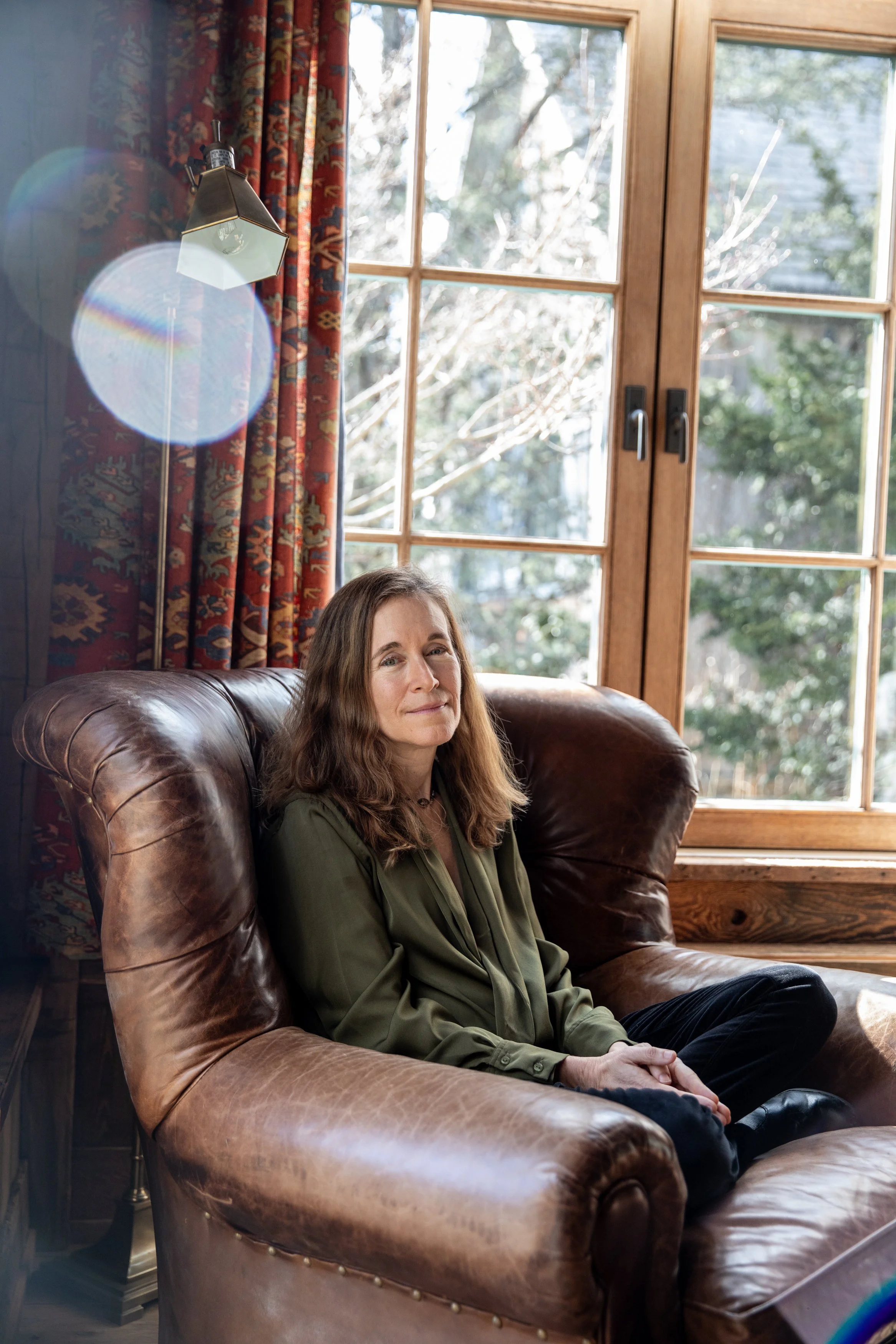 A woman with long brown hair sitting on a brown leather armchair in a cozy room near a large window with wooden frames and patterned curtains, with sunlight streaming in and trees visible outside.