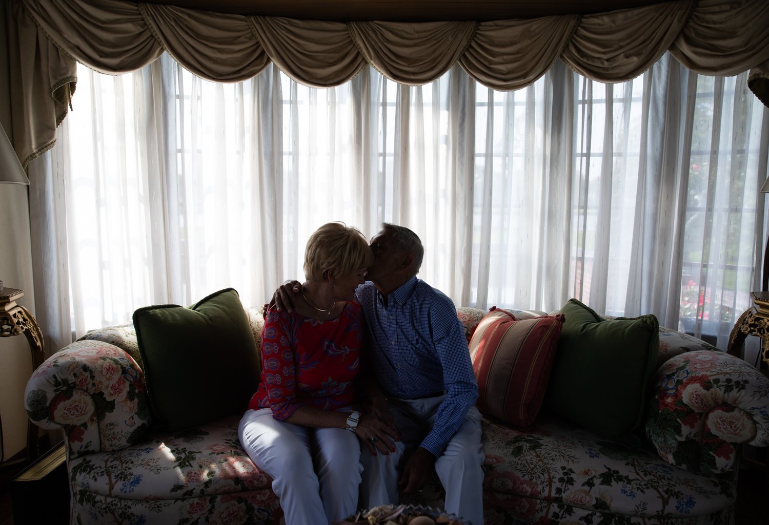 An elderly couple sitting close together on a floral-patterned couch in front of a large window with sheer curtains and valances, sharing an intimate moment.
