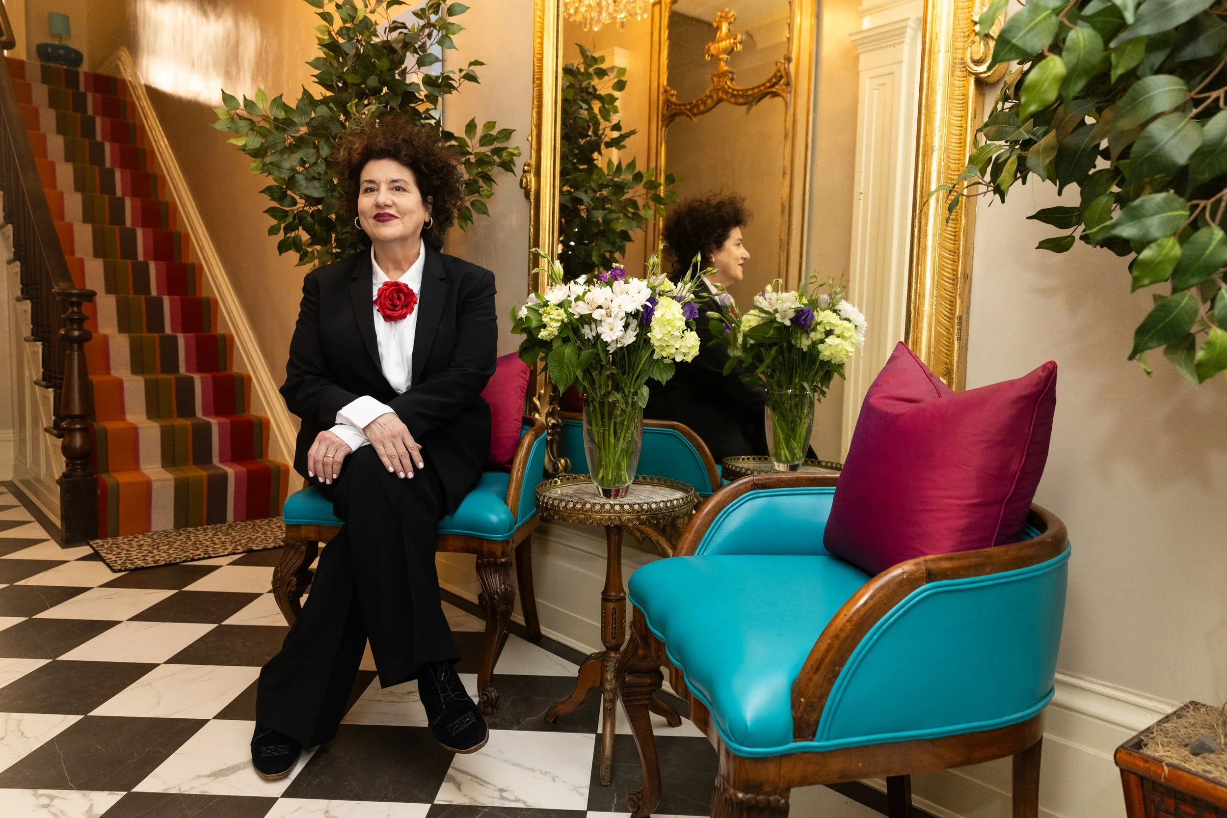 A woman with dark, curly hair and a red flower pin on her black blazer sits on a turquoise chair in an elegant lobby or sitting area. She has her arms crossed and is smiling slightly. Behind her, there are vibrant floral arrangements on a small round table, a gold-framed mirror reflecting part of her profile, ornate gold detailing on the mirror, and a staircase with a multi-colored striped carpet. The area features a checkered black-and-white floor and decorative greenery.