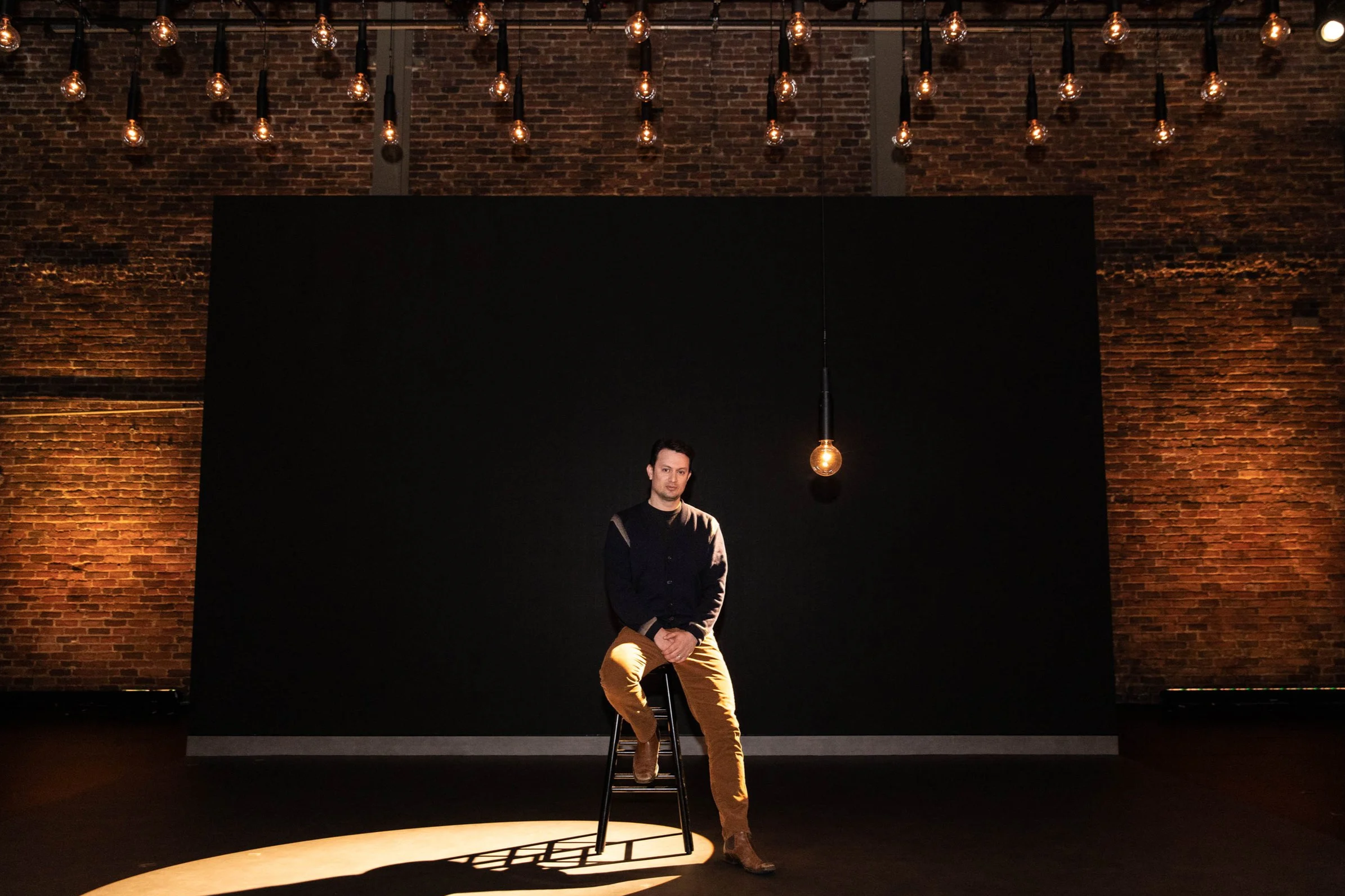 A man is sitting on a stool on a stage with a black background, in front of a brick wall with hanging light bulbs. The stage is illuminated by a spotlight, and the man is wearing khaki pants and a navy blue shirt.
