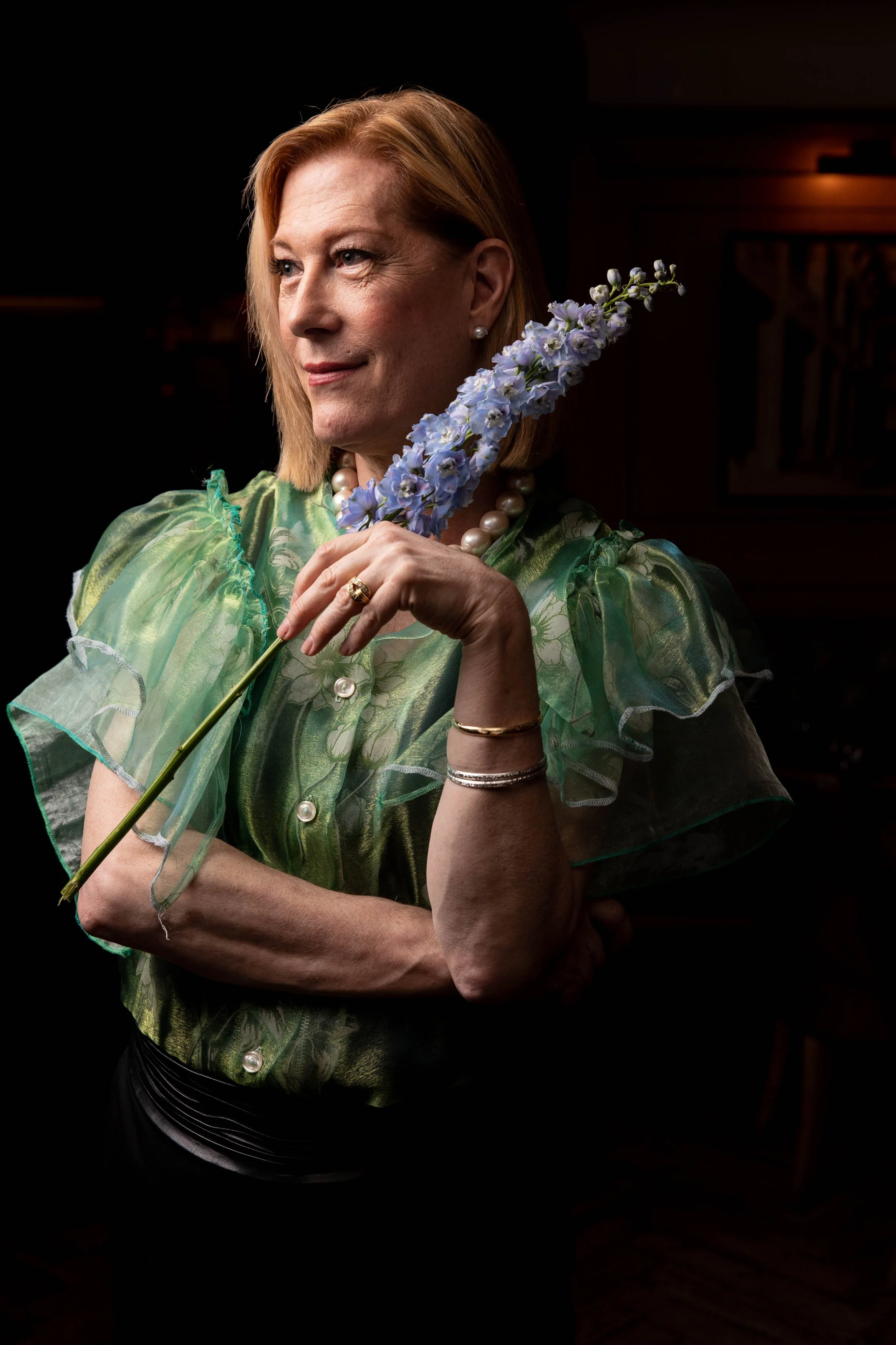 A woman with short blonde hair wearing a green ruffled blouse and pearl jewelry, holding a tall purple flower, with a dark background.