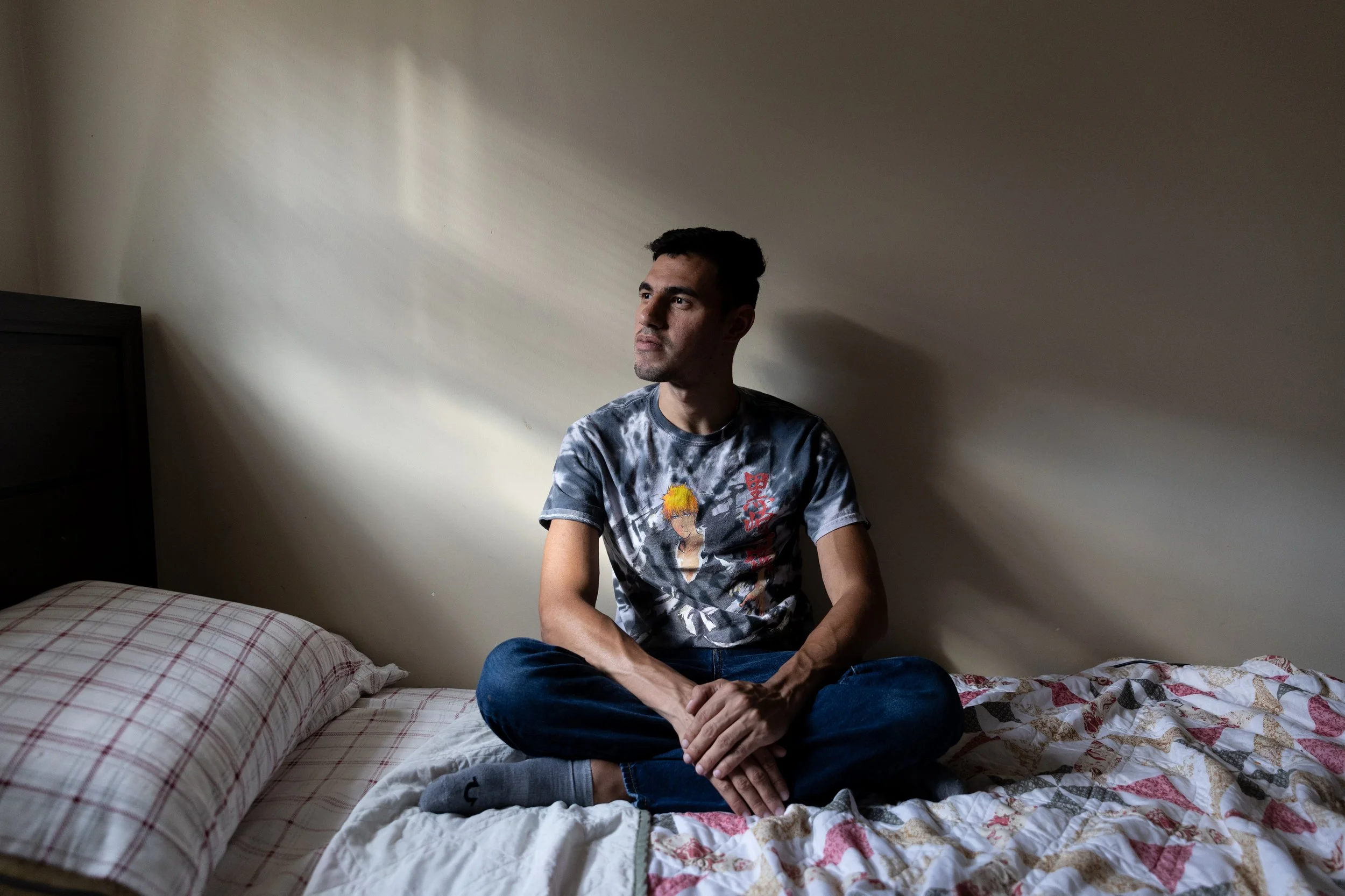 A young man with dark hair is sitting cross-legged on a bed, looking to his left with a contemplative expression. He is wearing a tie-dye t-shirt with an anime character on it and dark blue jeans. The room has plain beige walls, and sunlight is casting soft shadows.