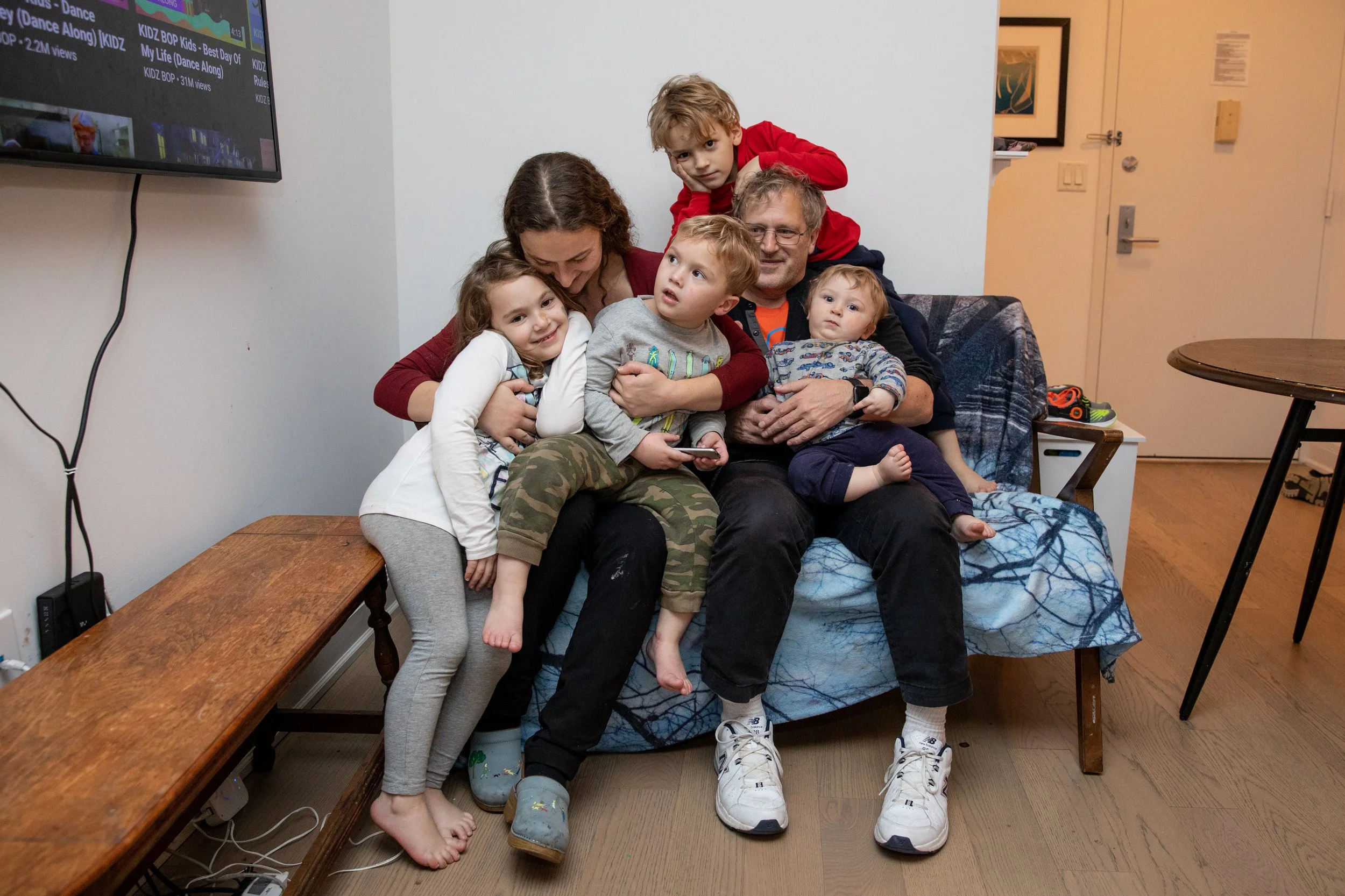 A family of six gathered closely together on a sofa in a living room, posing for a photo. The family includes an older man and woman, three young children, and a teenage boy. They are all smiling or looking at the camera, with one girl hugging the older man and a boy standing behind the sofa. The room has a wall with artwork, a flat-screen TV showing colorful content, and a small wooden table nearby.