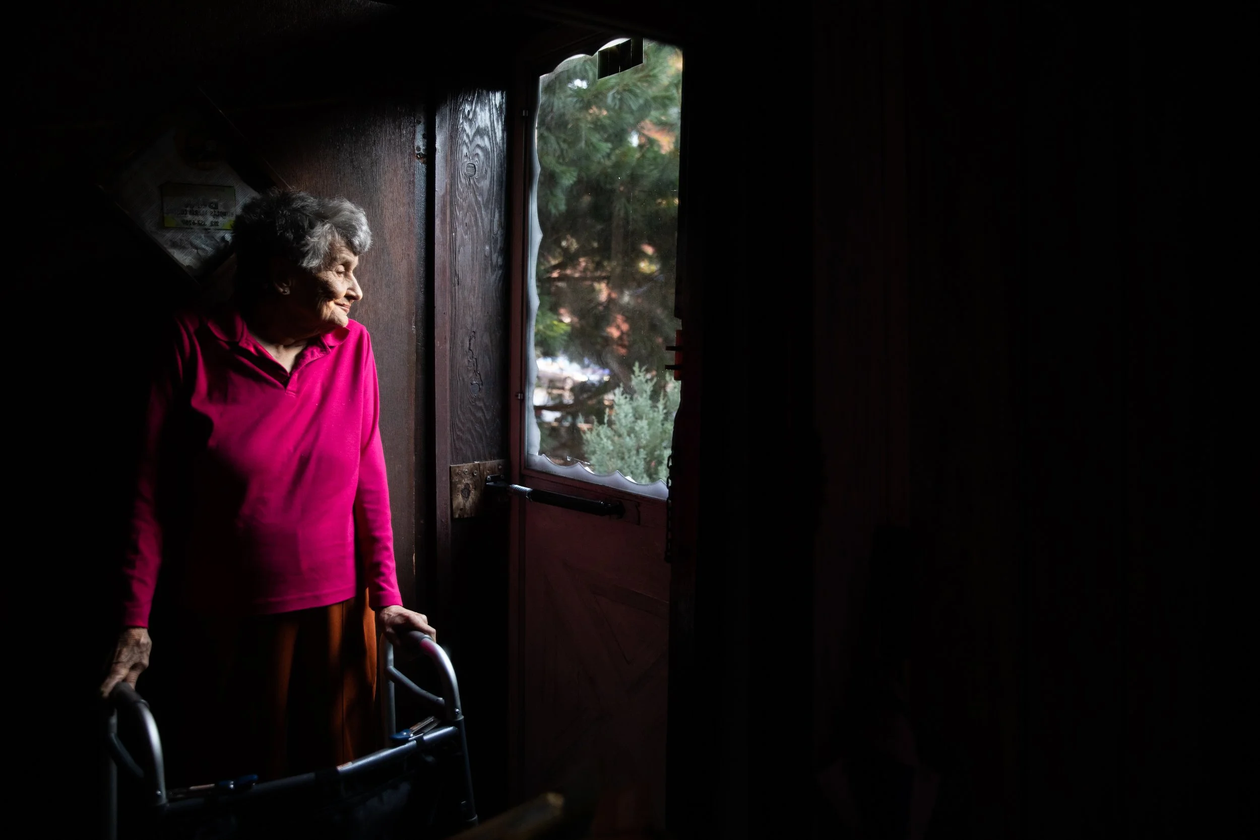 An elderly woman in a pink shirt standing with a walker by her side inside a dark room, looking outside through an open door. Sunlight illuminates her face and the greenery outside.