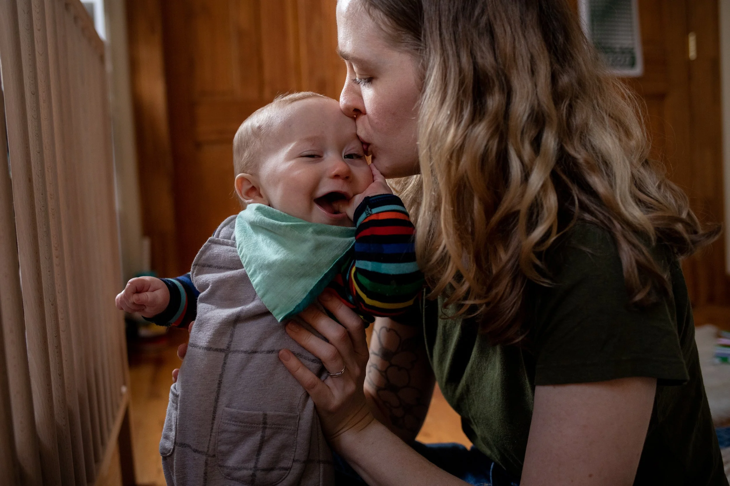 A woman with long, curly hair is giving a kiss on the forehead to a young boy with blonde hair, as he waves and smiles. They are in a warm wooden room.