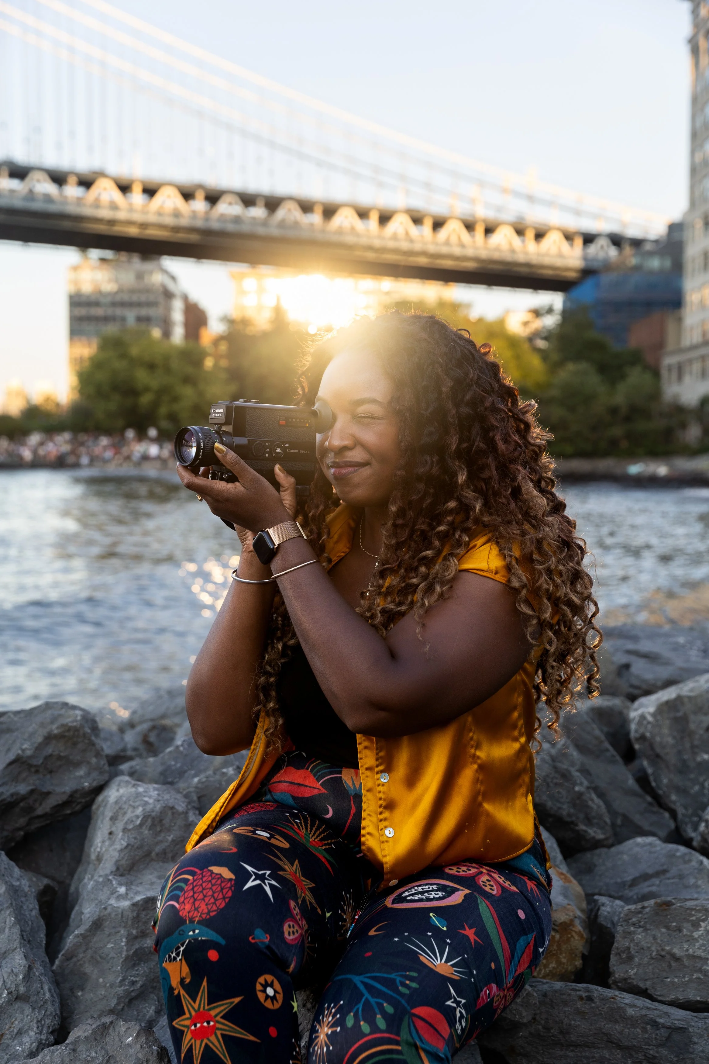 A woman with long curly hair, wearing a yellow jacket and colorful patterned pants, sitting on rocks near a river, holding a vintage camera to her eye, with the sun setting behind a bridge in the background.