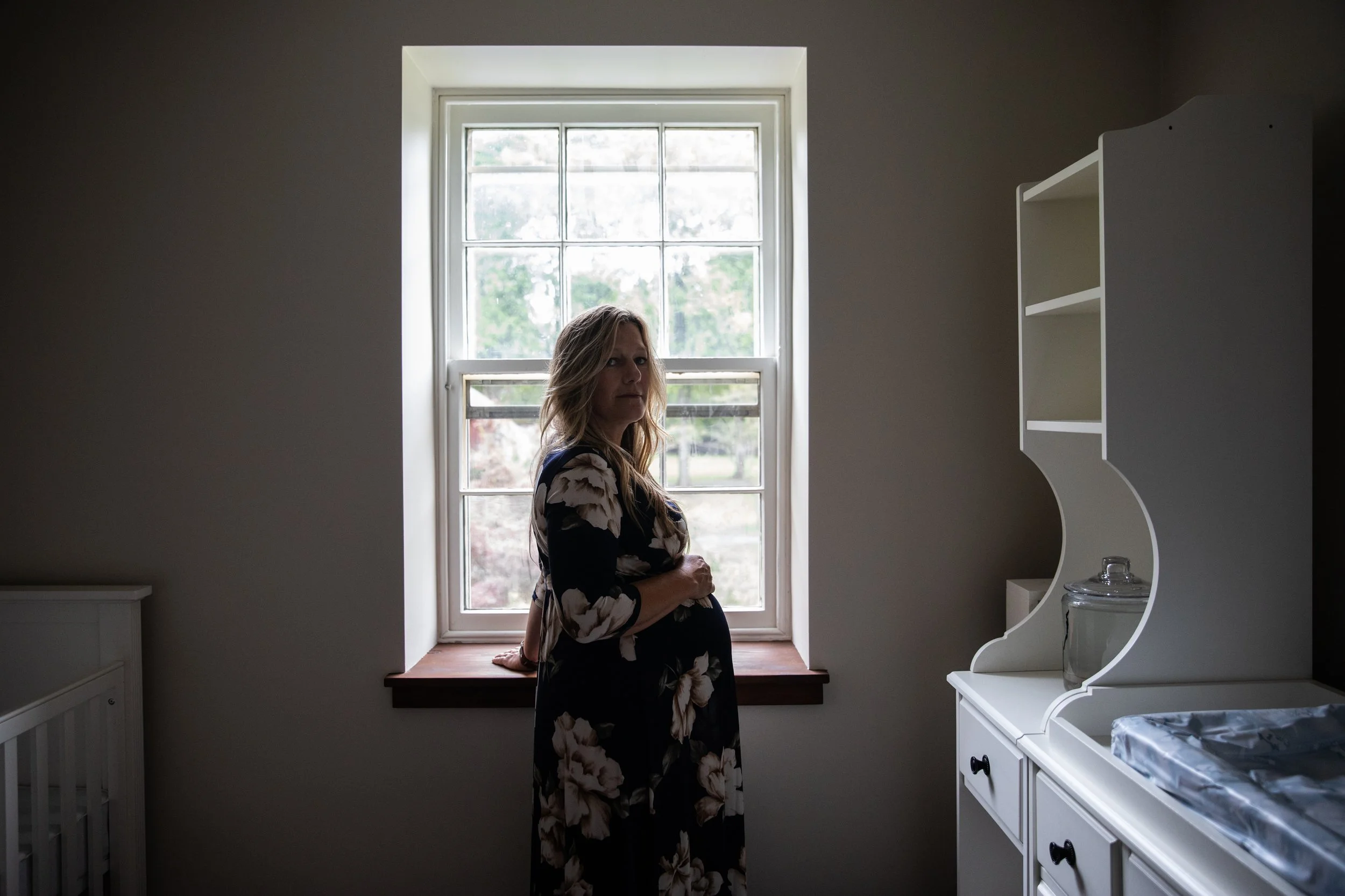 A woman with blonde hair wearing a black floral dress sitting on a windowsill in a room with beige walls, white furniture, and a window showing outside greenery.