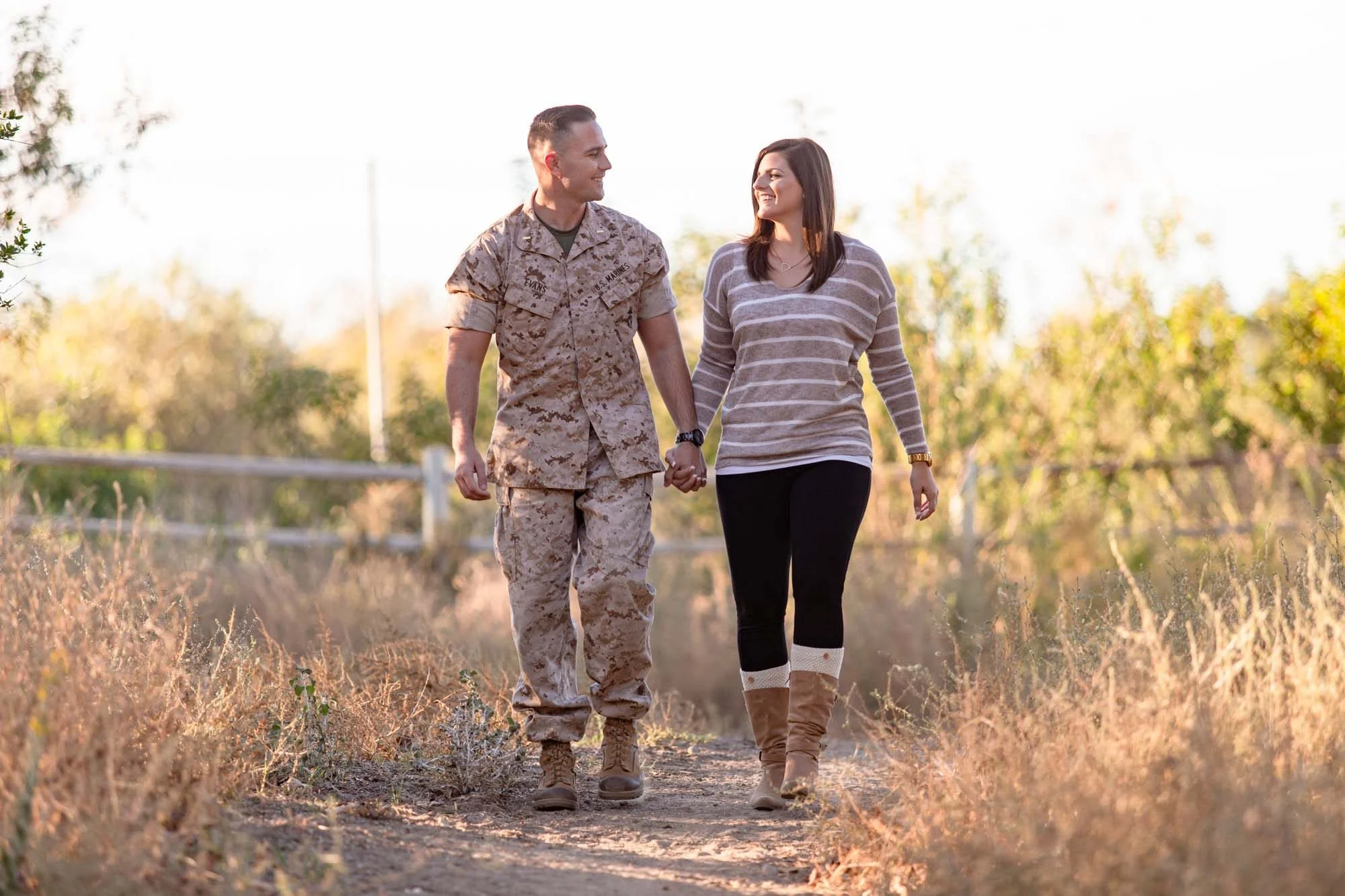 U.S. Marine in uniform walking hand in hand with his fiancée along a sunlit rural path during their engagement session