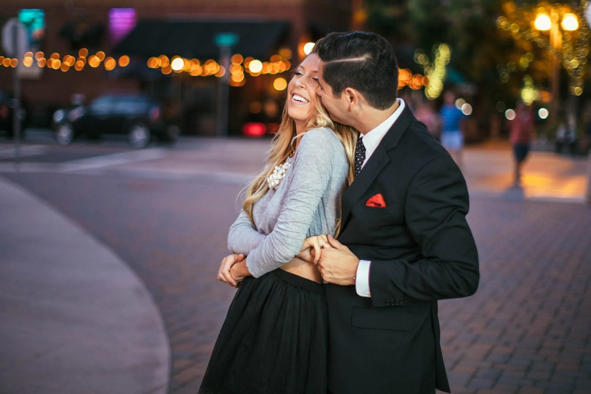 Engaged couple laughing and embracing on a downtown Riverside street at night with golden string lights glowing softly behind them