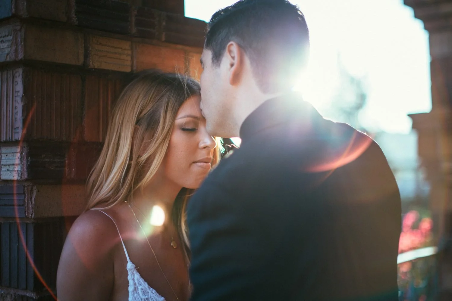 Intimate close-up of couple nearly forehead to forehead against a Mission Inn brick wall with soft golden sunflare