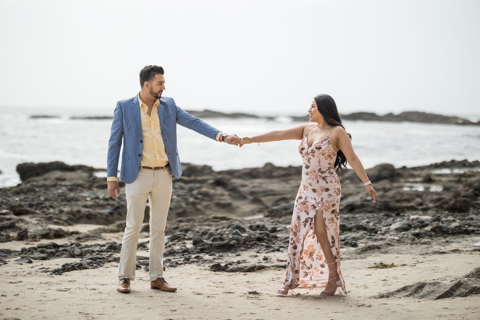 Well-dressed couple holding hands on the rocky coast at Laguna Beach, woman in a floral dress with the Pacific Ocean behind them