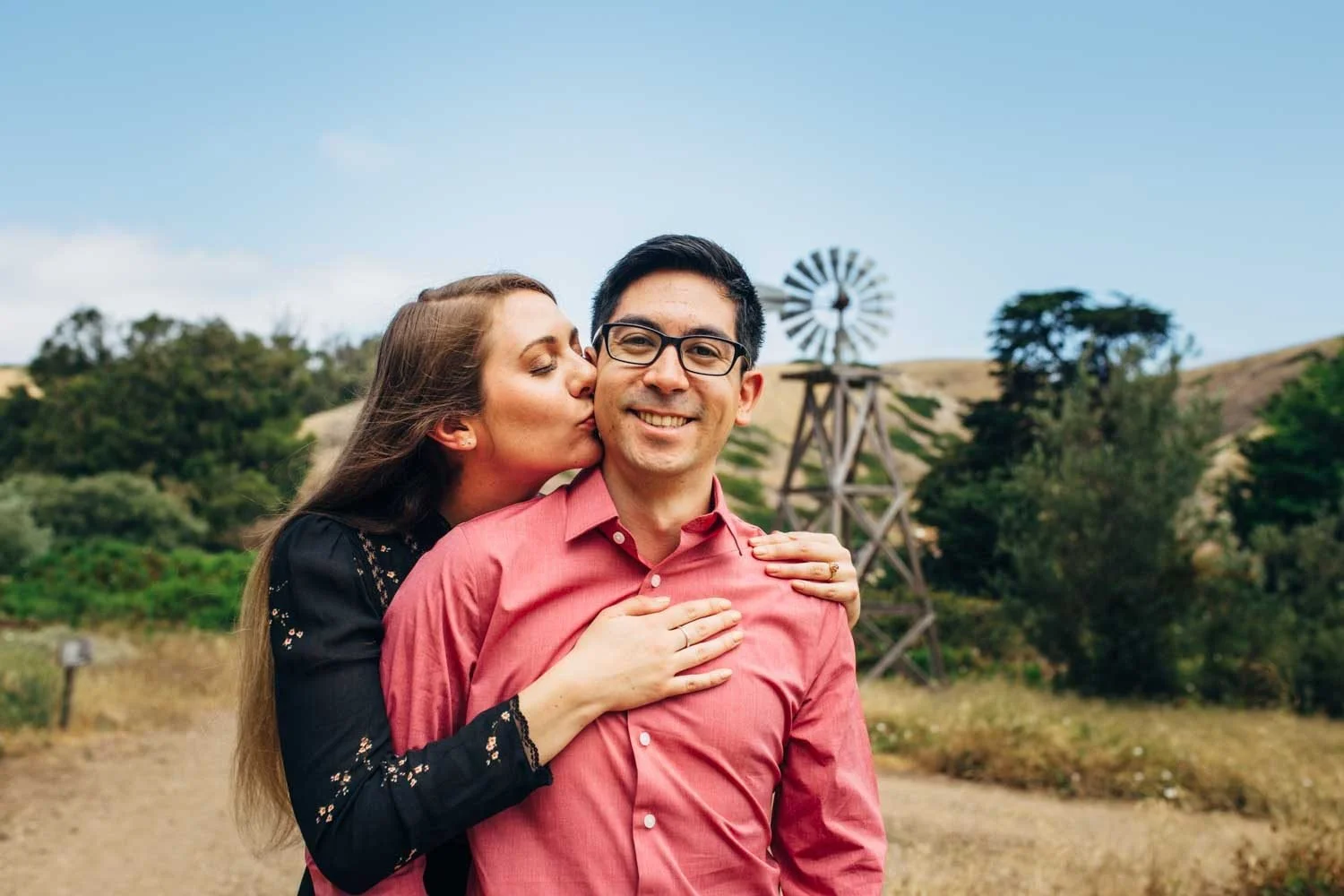 Woman kissing her partner's cheek during a Santa Cruz Island engagement session, with a rustic windmill and golden rolling hills behind them
