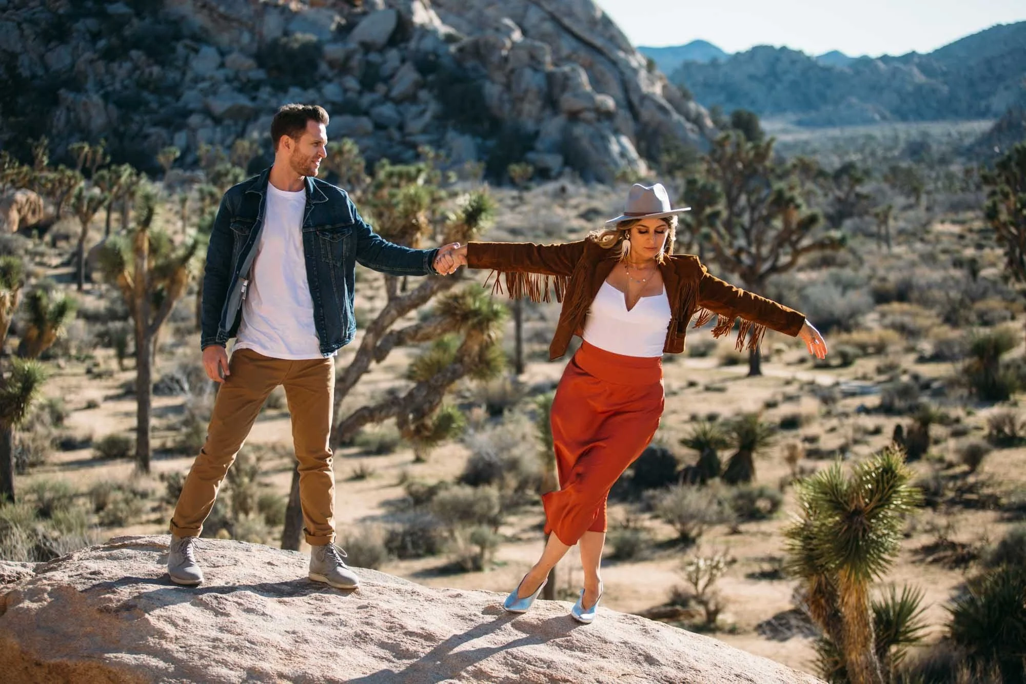 Boho engaged couple holding hands and walking across granite boulders surrounded by Joshua trees and desert scrub