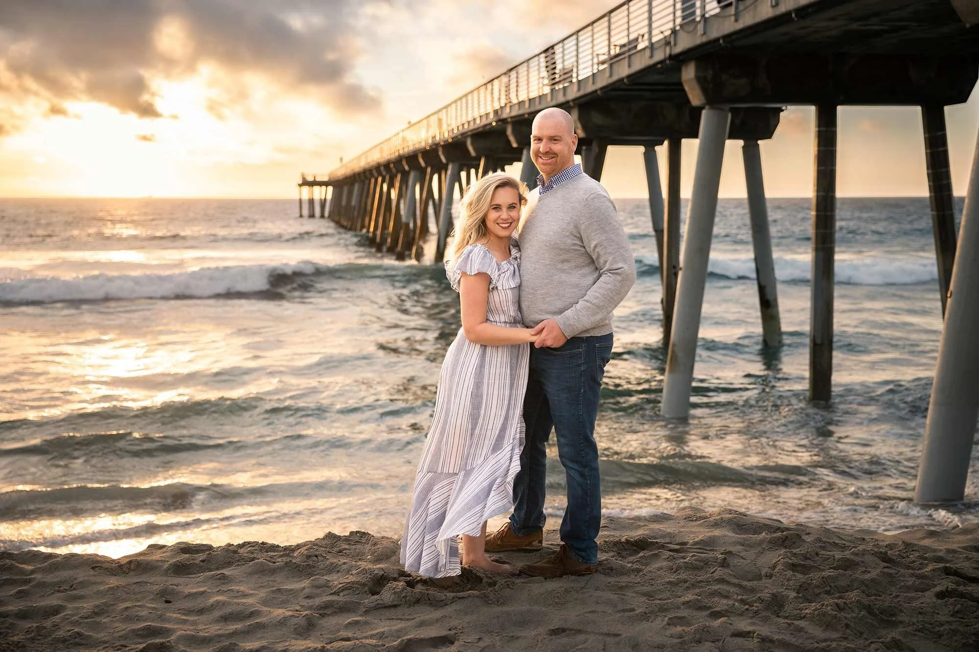 Smiling engaged couple standing arm in arm beneath Hermosa Beach pier during a warm golden sunset