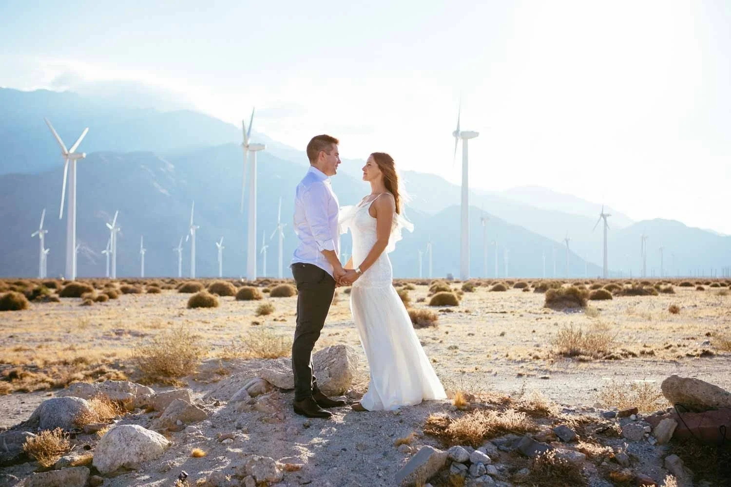 Engaged couple standing face to face holding hands in a Palm Springs desert with wind turbines and mountains filling the horizon