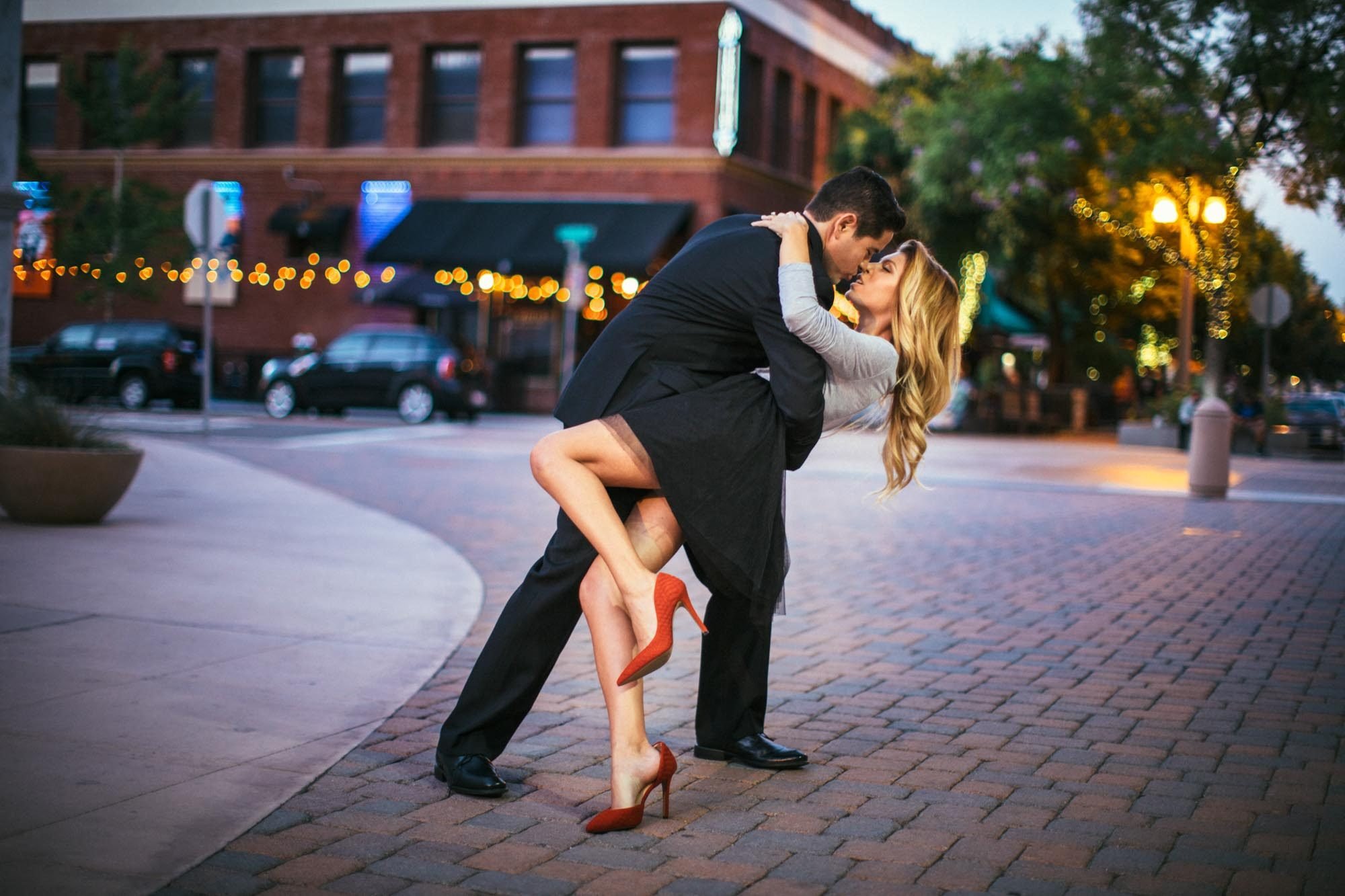 Couple sharing a dramatic dip kiss on a downtown Riverside brick plaza at dusk with warm string light bokeh glowing behind them