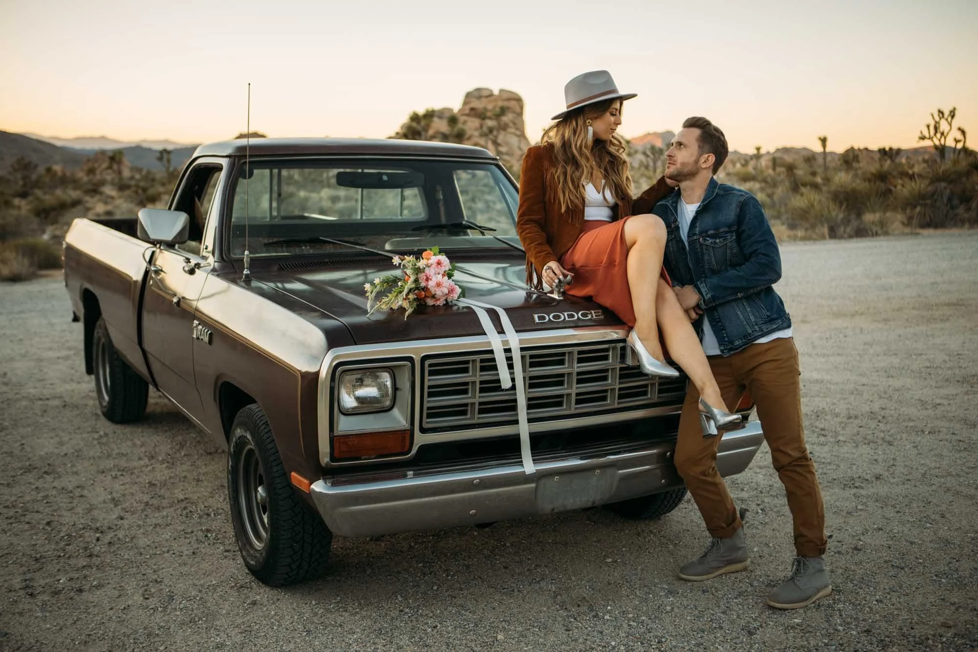 Engaged couple with a vintage Dodge truck at Joshua Tree sunset, woman seated on the hood with wildflower bouquet