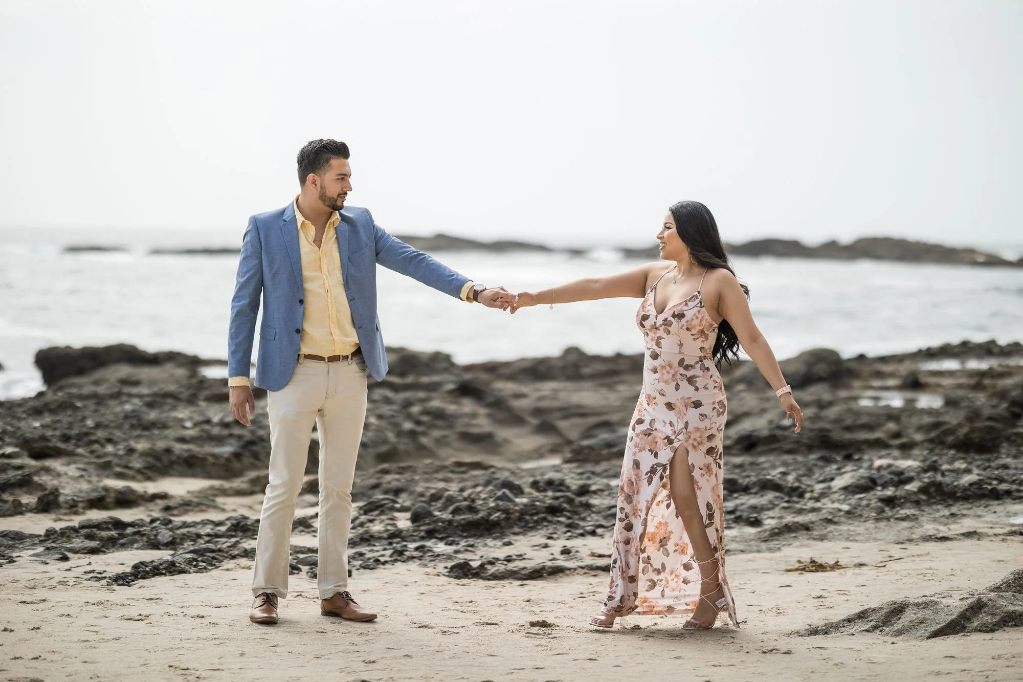 Well-dressed couple holding hands on a rocky Southern California beach, woman in a floral dress with the gray ocean behind them