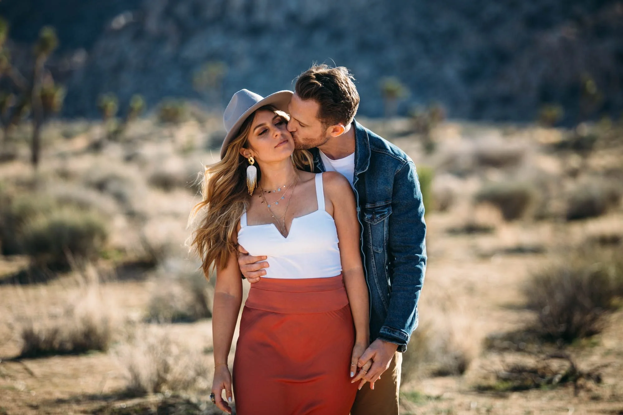 Man kissing woman's cheek during a Joshua Tree engagement session, warm desert light and boulders softly blurred behind them