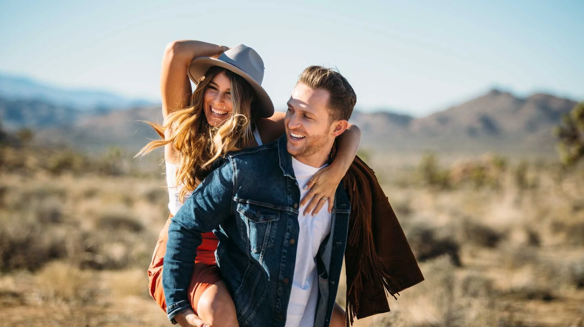 Laughing boho couple during a Joshua Tree engagement session, woman in wide-brim hat riding piggyback in the desert