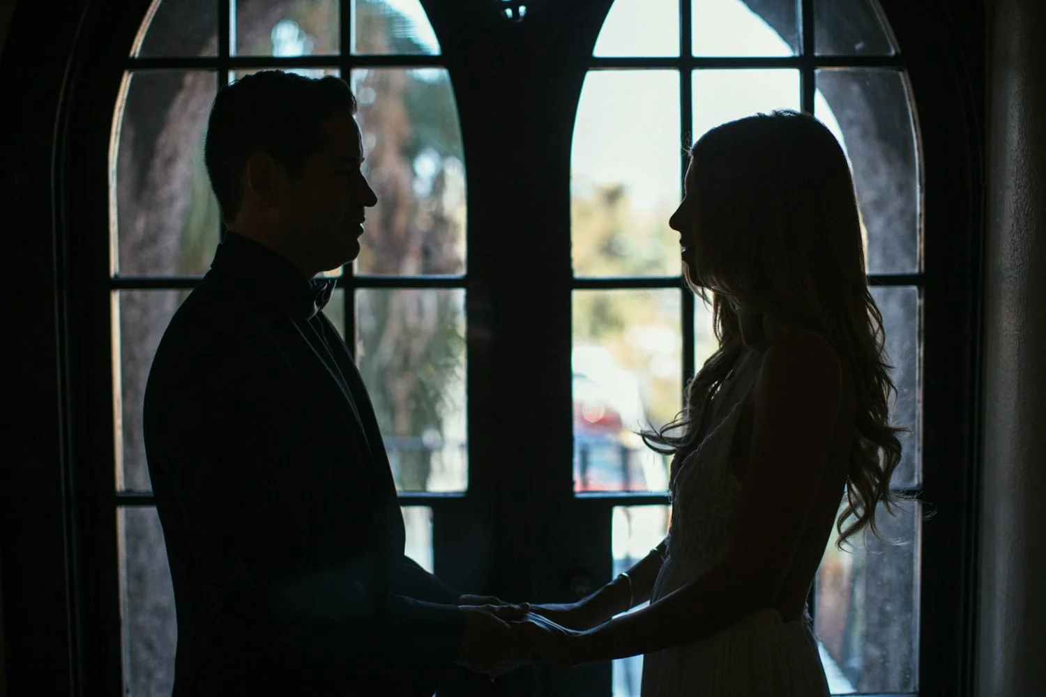 Engaged couple silhouetted face to face holding hands in front of Mission Inn's tall arched windows in dramatic low light