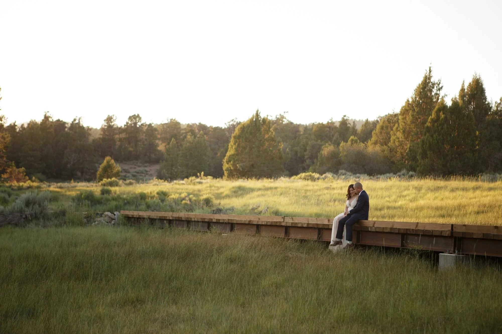 Couple sitting close together on a wooden bridge kissing at golden hour at Juniper Point in Big Bear Lake, California, surrounded by a glowing green meadow and pines