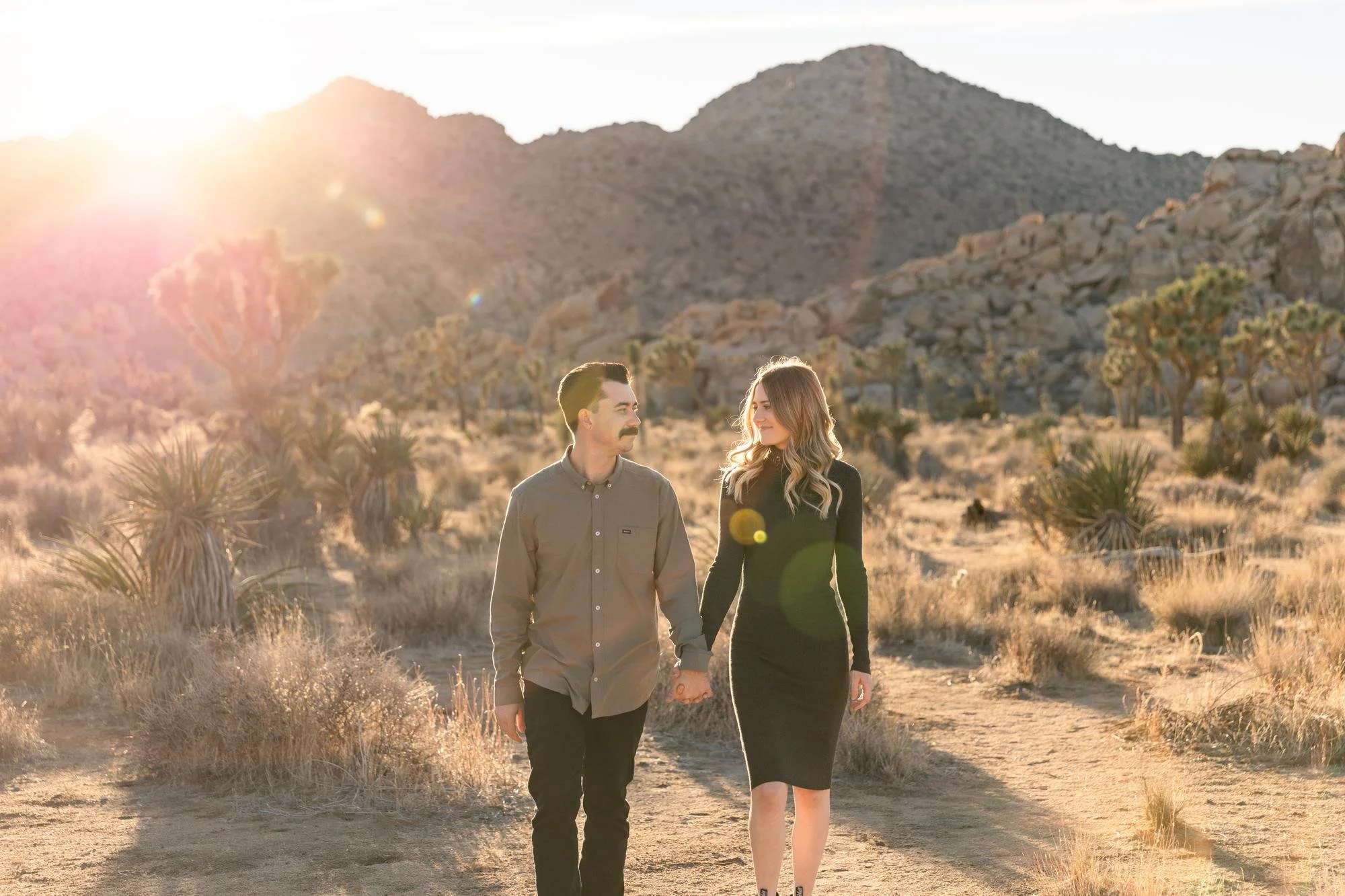 Couple walking hand in hand through Joshua Tree desert at golden hour with sun flare and rocky mountains behind them