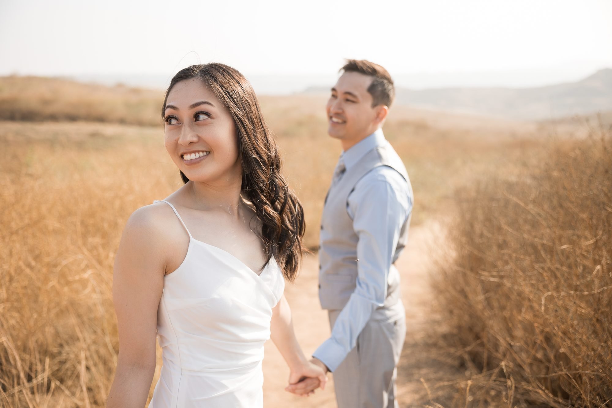 Engaged woman in white dress leading her partner by the hand through a golden California hillside field at sunset