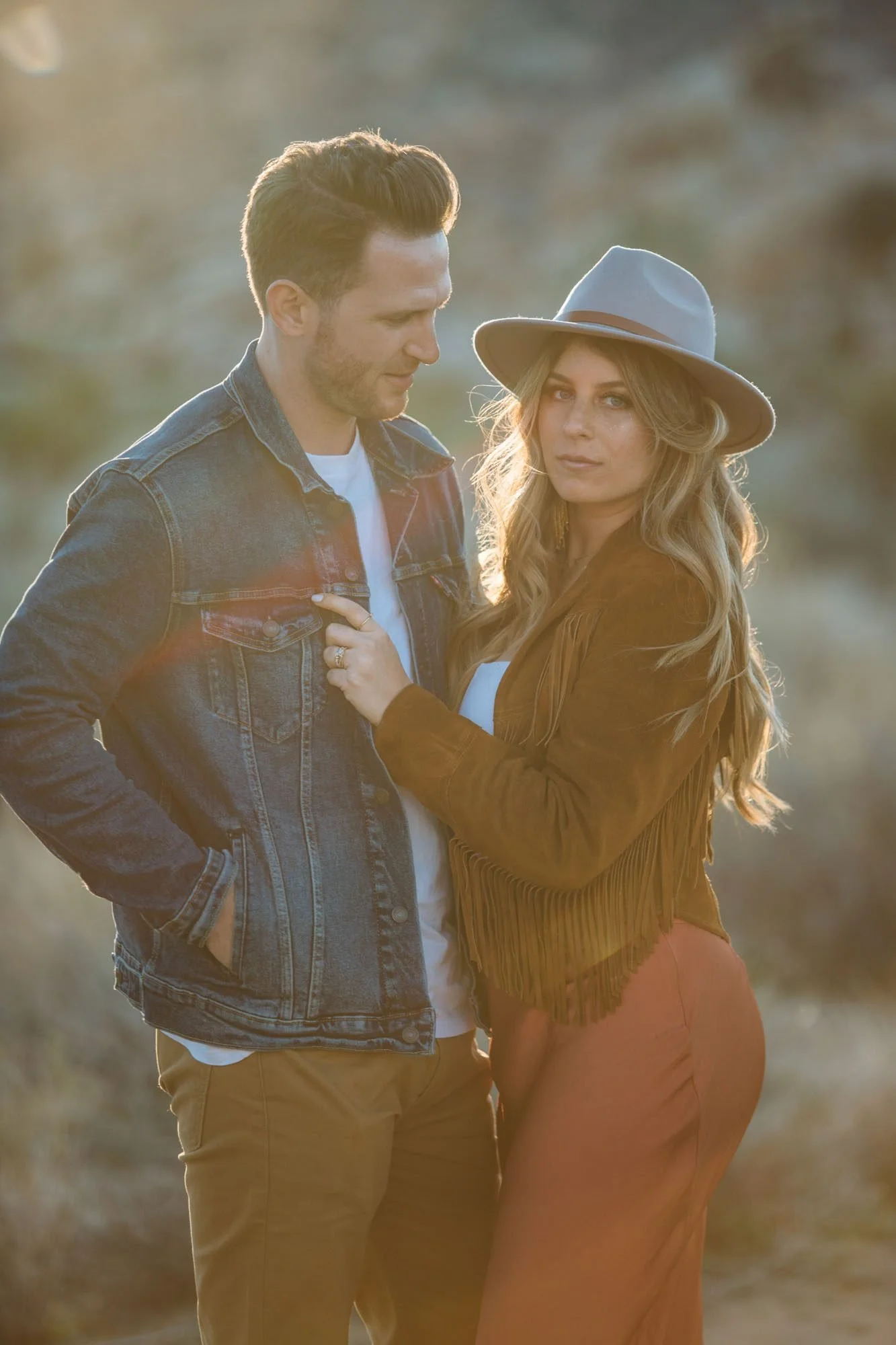 Boho engaged couple close-up in Joshua Tree golden hour light, woman in fringe jacket and wide-brim hat gazing softly