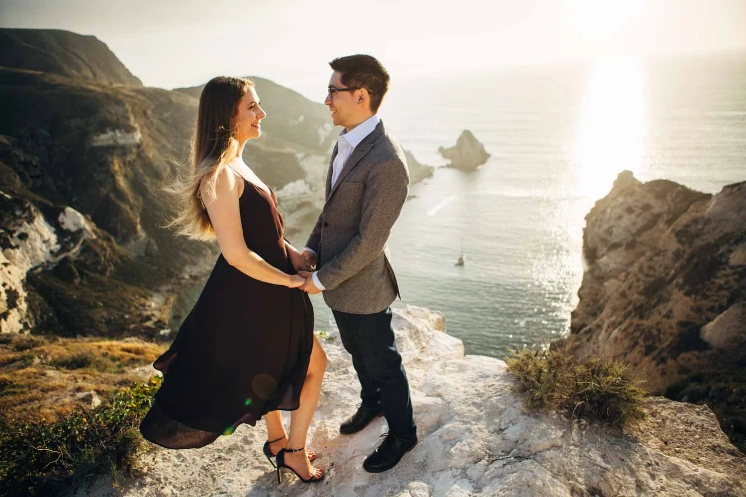 Engaged couple holding hands and smiling on a rocky cliff edge at Santa Cruz Island, Channel Islands National Park, with glittering ocean and sea stacks below