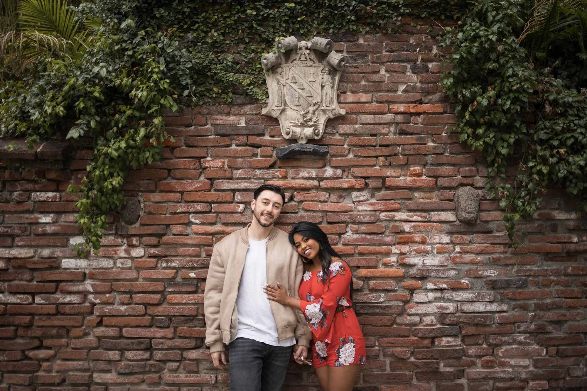 Smiling couple leaning against Mission Inn's ivy-draped brick wall beneath a carved stone crest during their Riverside engagement session