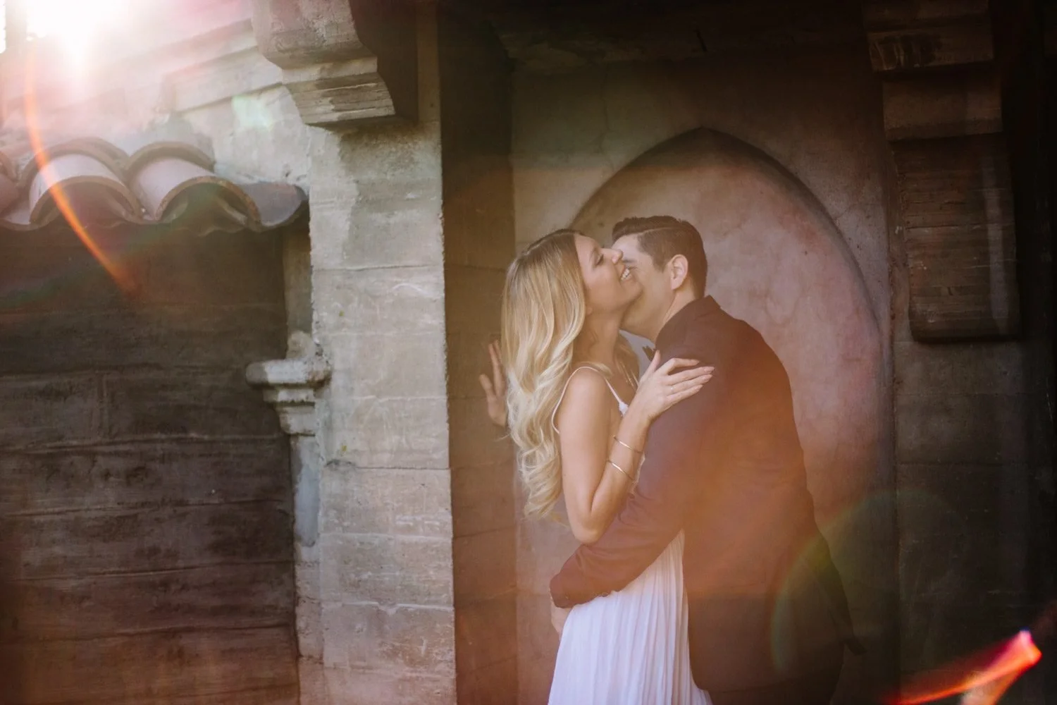 Couple kissing beneath a Mission Inn stone arch with terracotta roof tiles and a warm golden sunflare washing over them