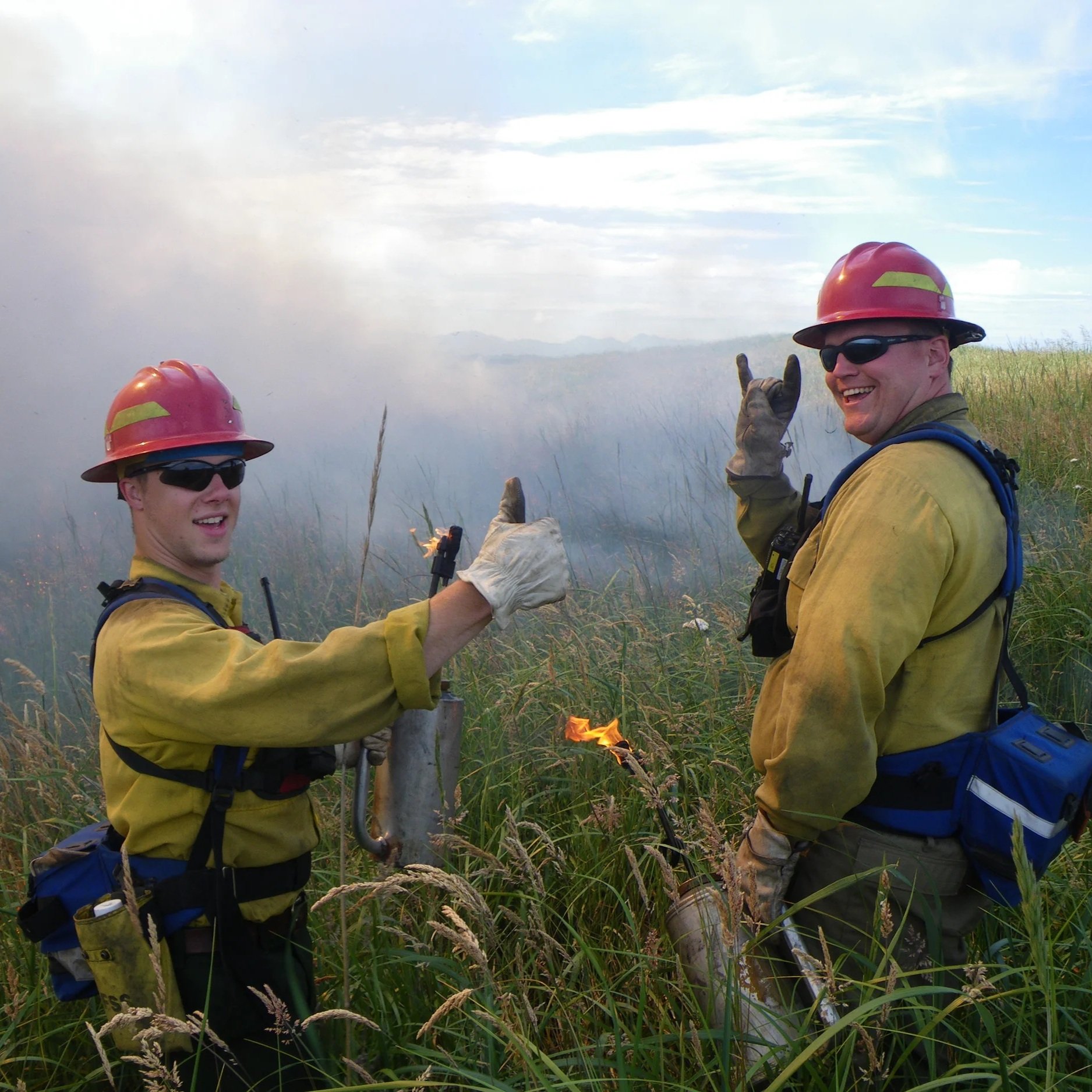 Two firefighters in yellow protective gear and red helmets stand in tall grass, holding a flaming torch and giving a thumbs-up, with smoke rising in the background.